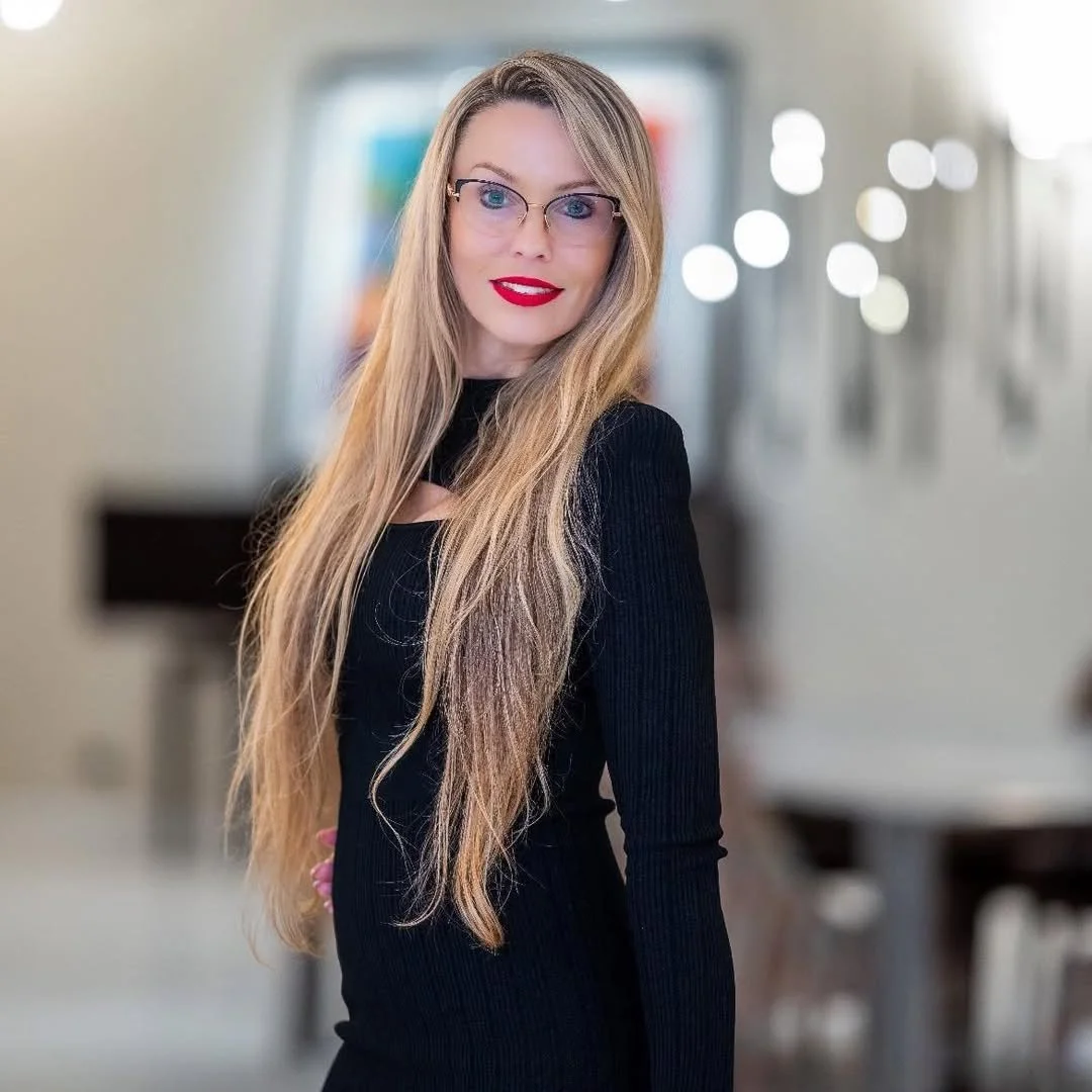 A young woman with long blonde hair, wearing glasses and a black top, standing in a modern indoor setting with blurred background lights.