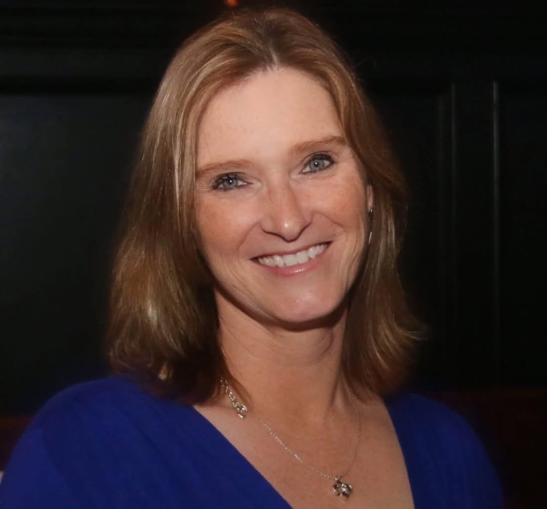A woman with shoulder-length reddish-brown hair, wearing a blue top and a necklace, smiling at the camera.