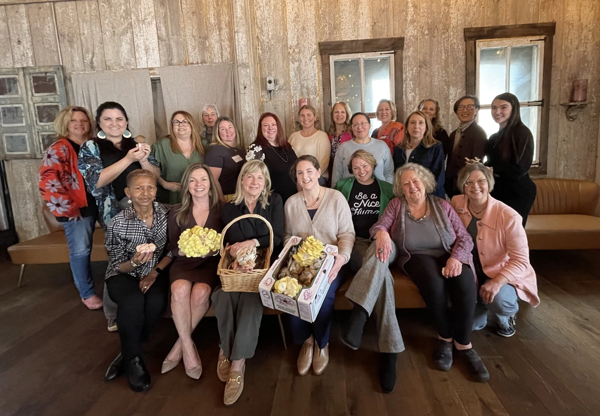 Group of women gathered indoors, some holding flowers and baked goods, smiling for a photo in a rustic room with wooden walls and windows.