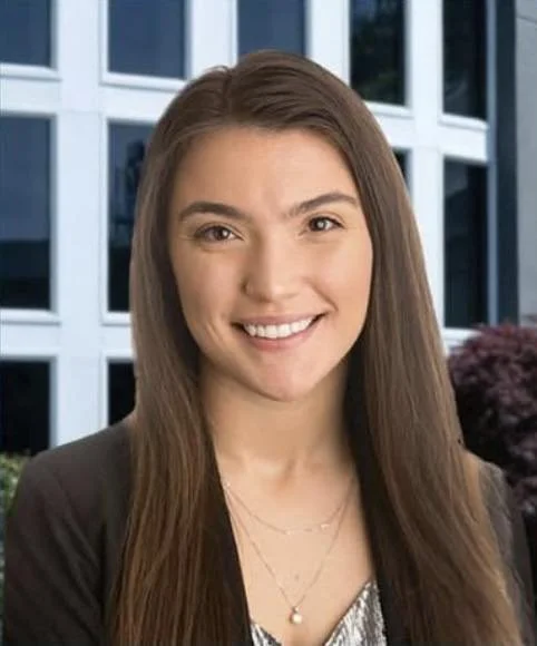 A young woman with long brown hair smiling, standing outdoors in front of a modern glass building.