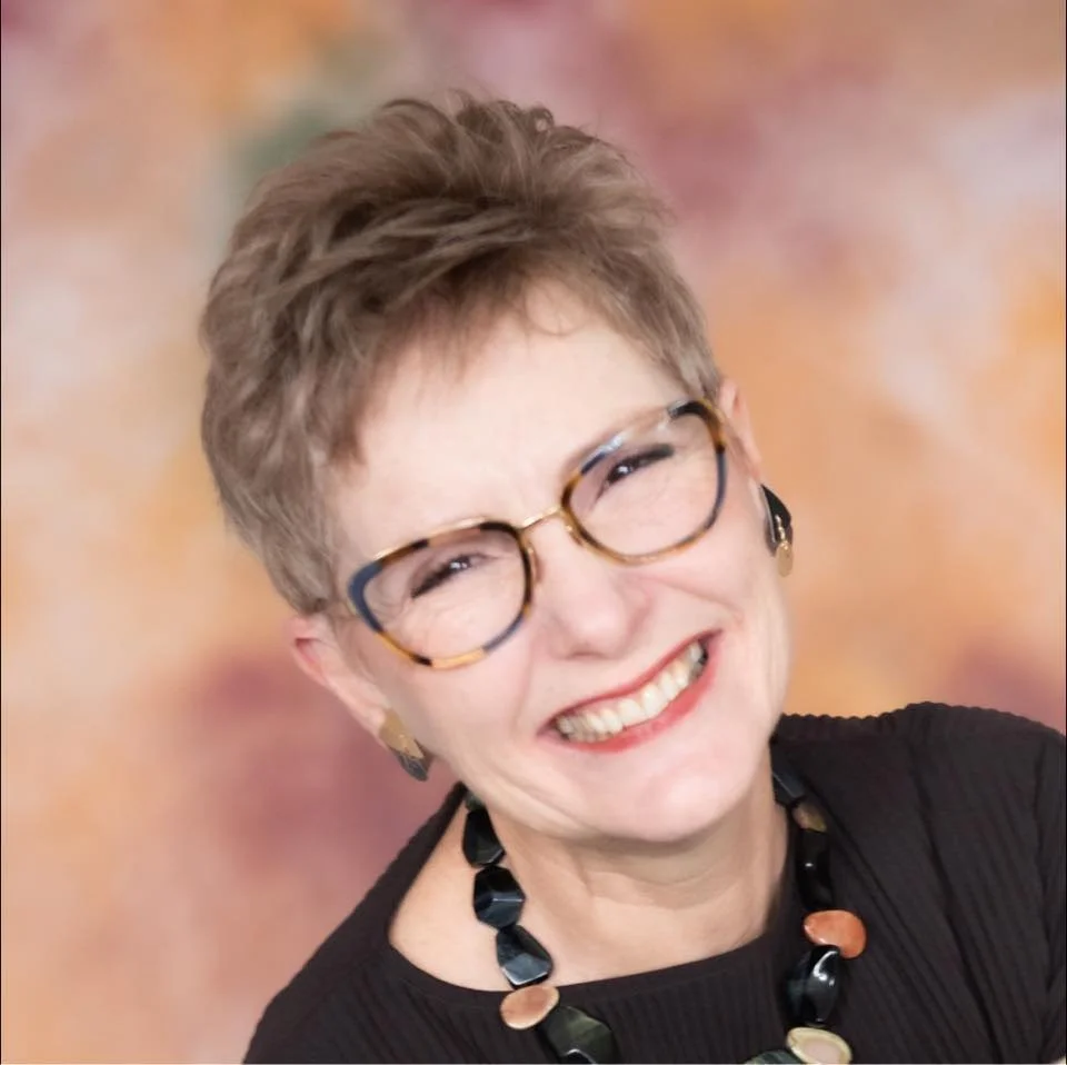 A woman with short, tousled brown hair wearing glasses with tortoise shell frames, a black top, and a statement necklace with large black and beige beads, smiling brightly against a colorful, blurred background.