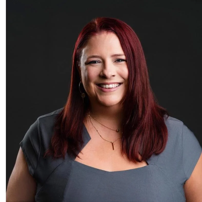 A woman with red hair smiling, wearing a gray top and layered necklaces, standing against a dark background.