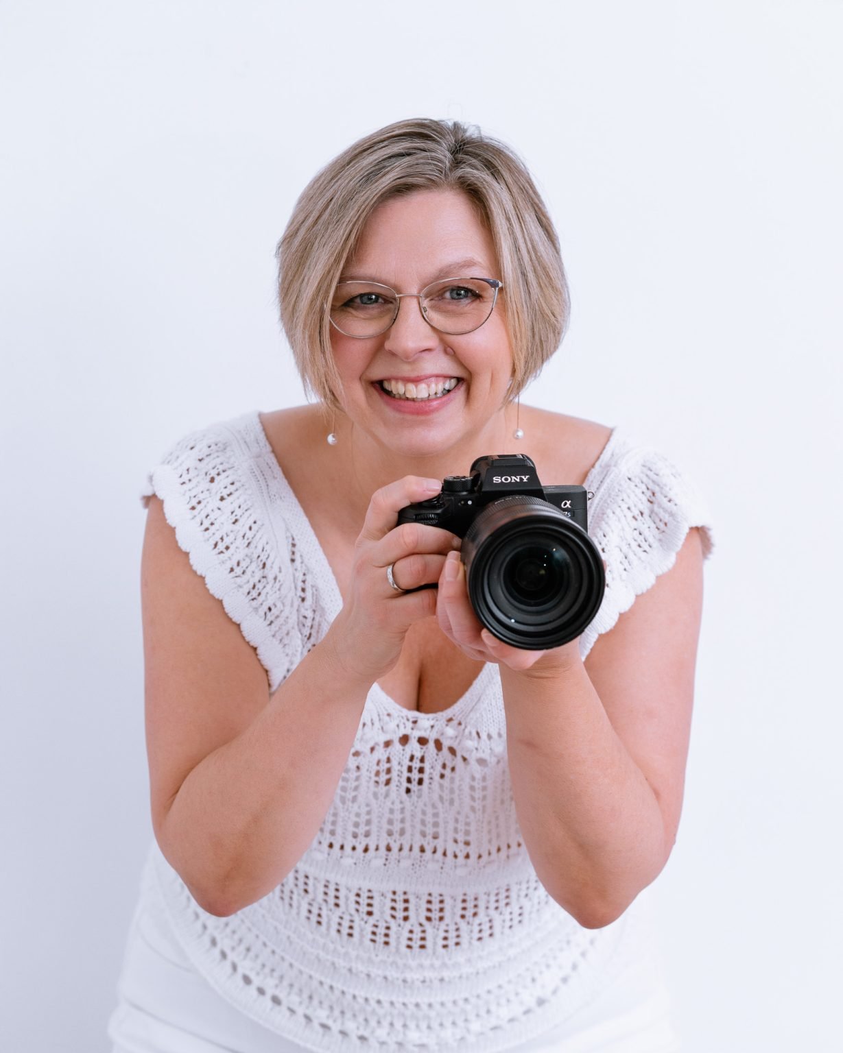 A smiling woman with short blonde hair and glasses, holding a Sony camera, wearing a white crocheted top, against a plain white background.