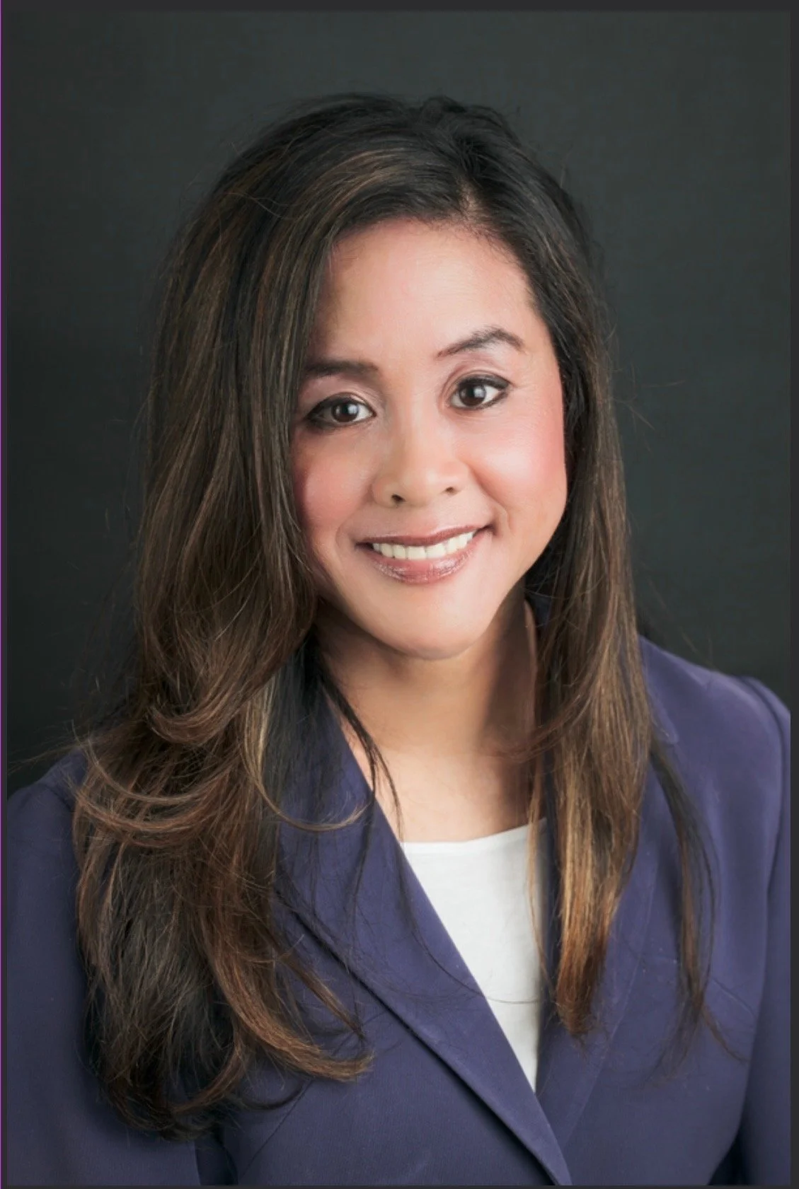 A professional woman with long, wavy brown hair and a light smile, wearing a dark blazer over a white top, posing against a dark background.