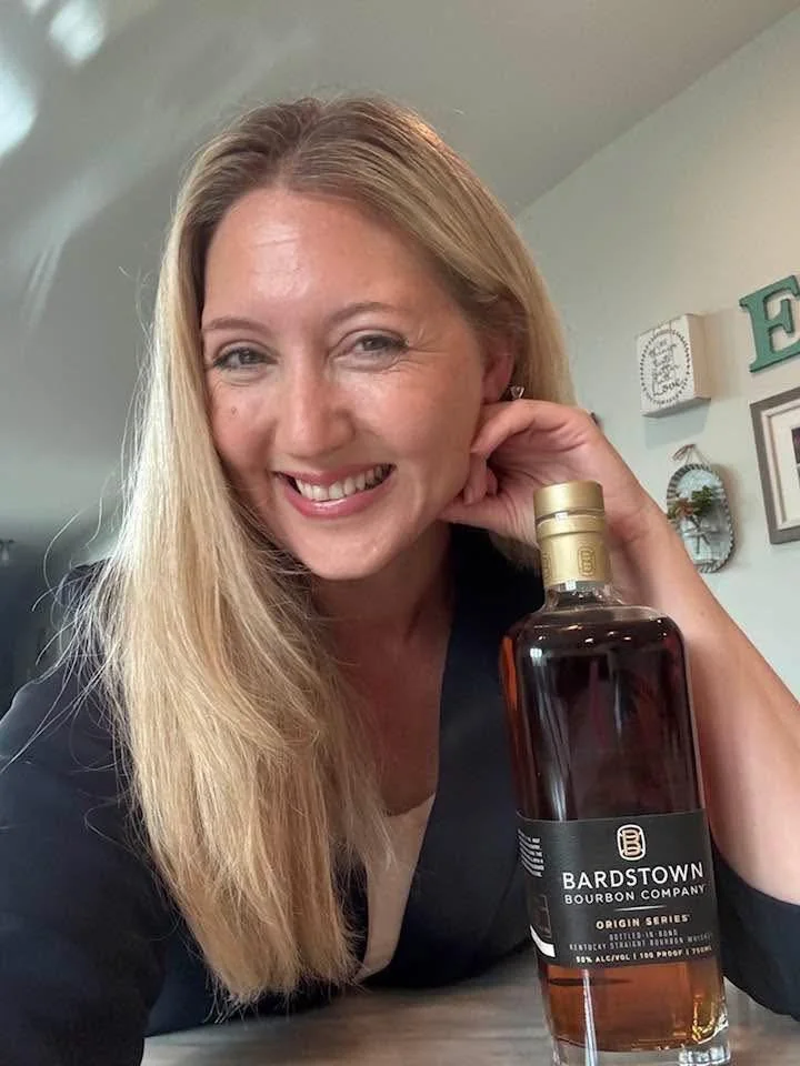 A woman with long blonde hair smiling and holding a bottle of Bardstown bourbon company whiskey.