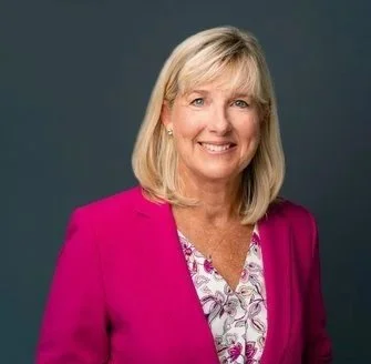 Portrait of a smiling woman with blonde hair, wearing a pink blazer and a floral blouse, against a dark background.