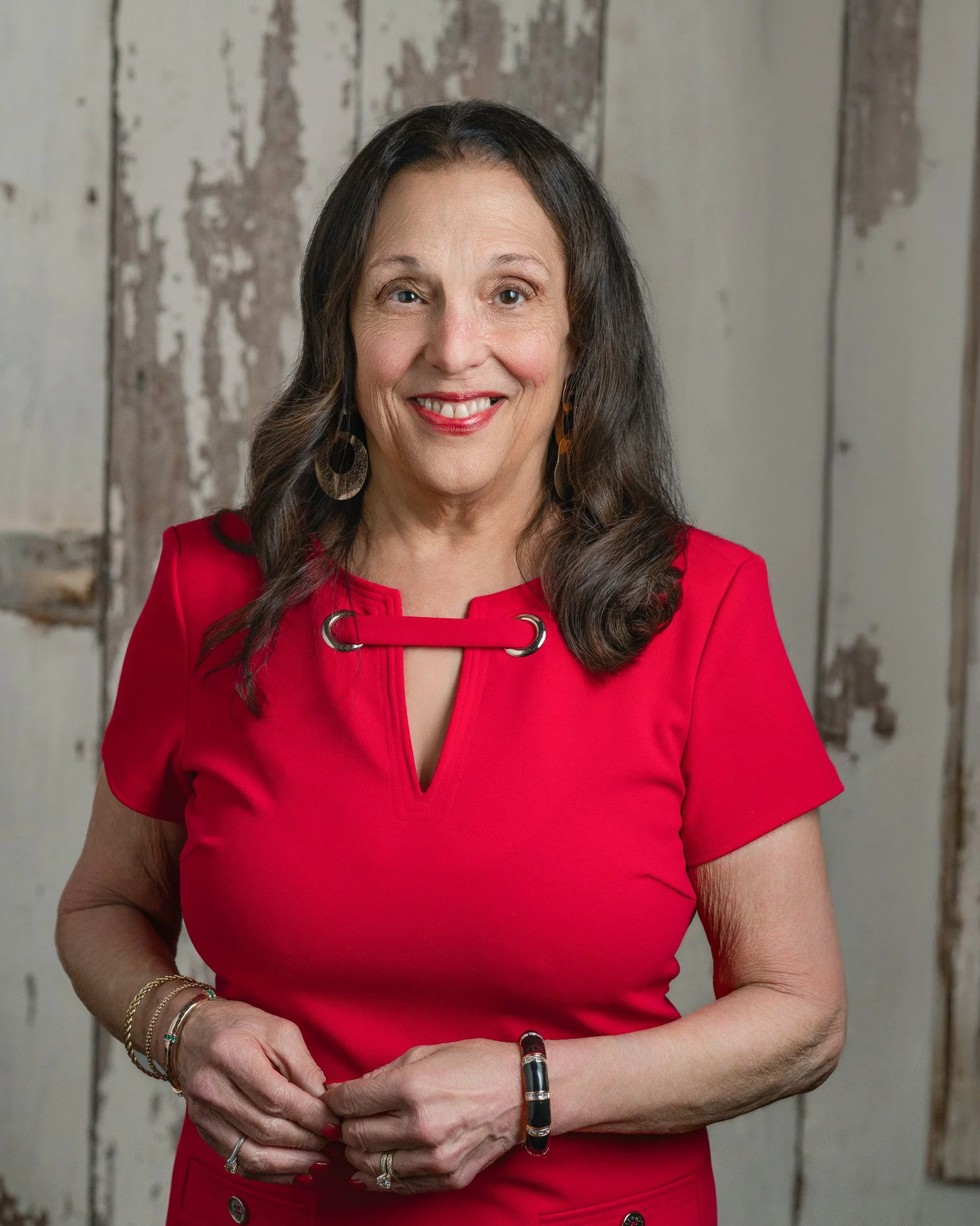 A smiling woman with glasses and long dark hair, wearing a blue sleeveless top and white shorts, sitting on a chair against a plain gray background.