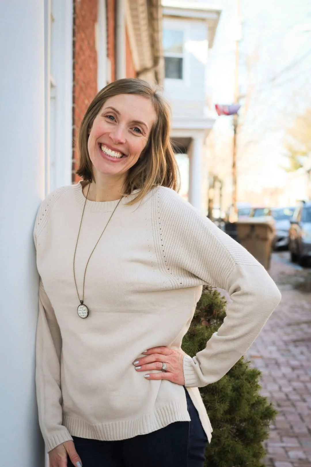 A woman with shoulder-length brown hair smiles widely while leaning against a white wall on a city sidewalk in autumn, wearing a cream sweater and a long necklace.