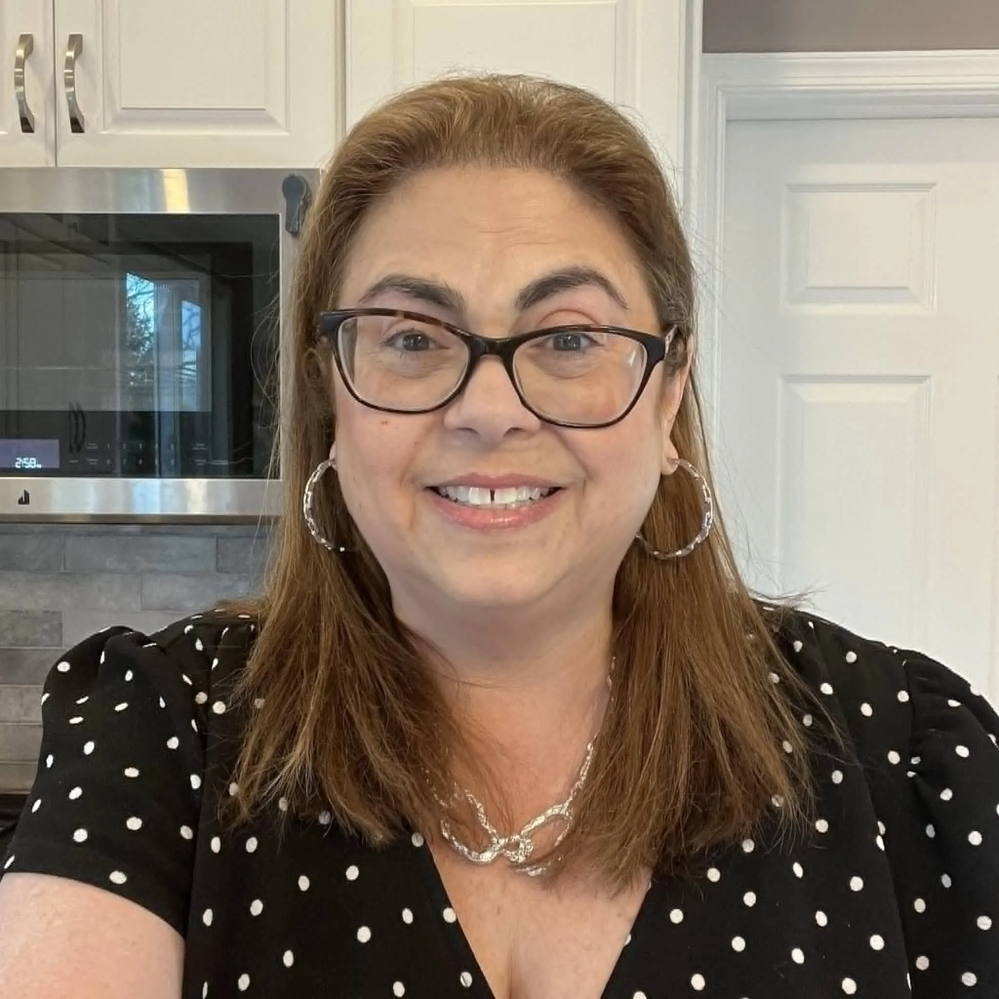 A woman wearing glasses and earrings smiling, sitting indoors with a window behind her showing a yard with trees and a patio with outdoor furniture.
