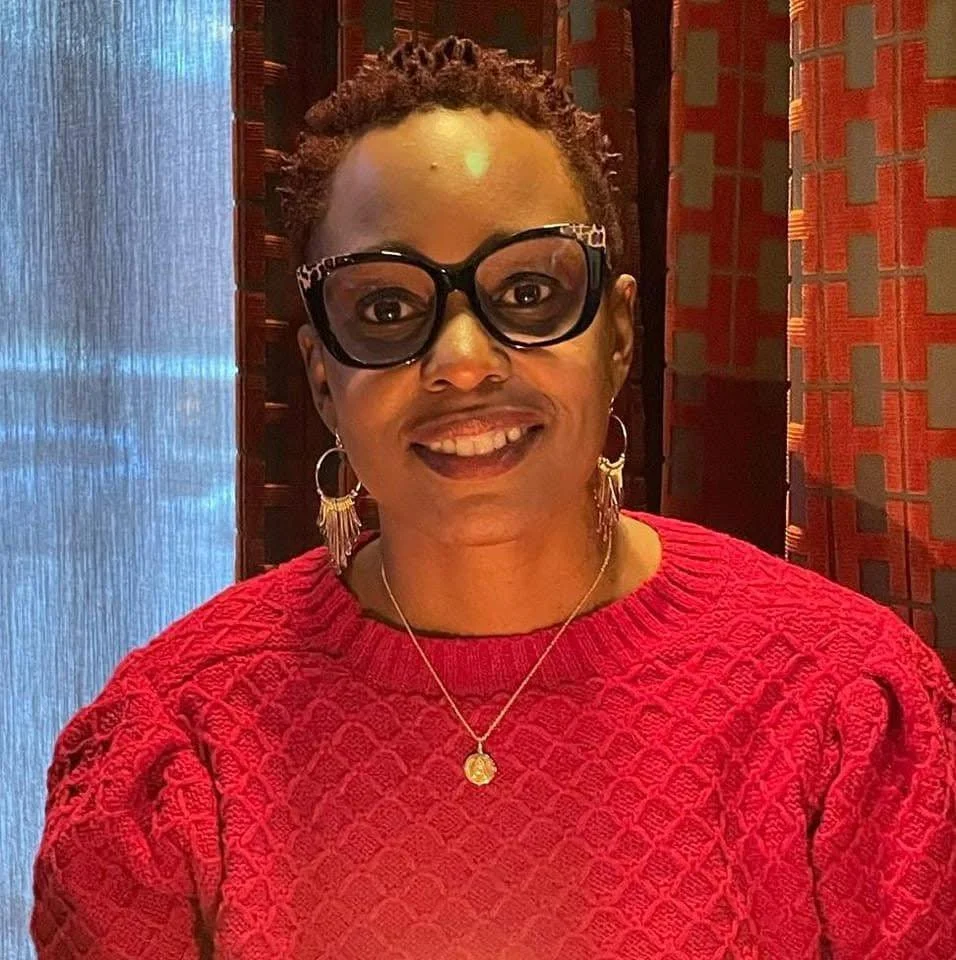A woman with short curly hair, wearing large black cat-eye glasses, silver hoop earrings with fringe, a gold necklace, and a red textured sweater. She is smiling in front of a patterned background.