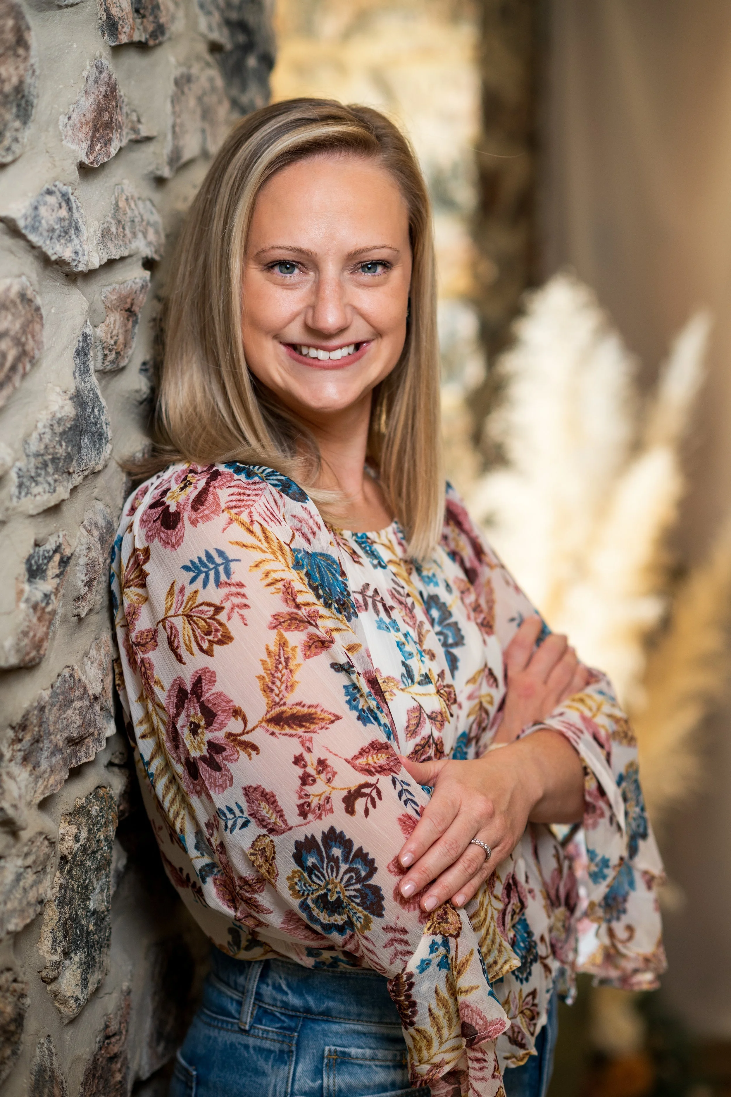 A woman with shoulder-length blonde hair smiling, leaning against a stone wall with crossed arms, outdoors.