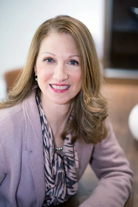 A woman with long, wavy light brown hair and fair skin smiling, wearing a light pink blazer and a patterned blouse, sitting indoors with a blurred background.