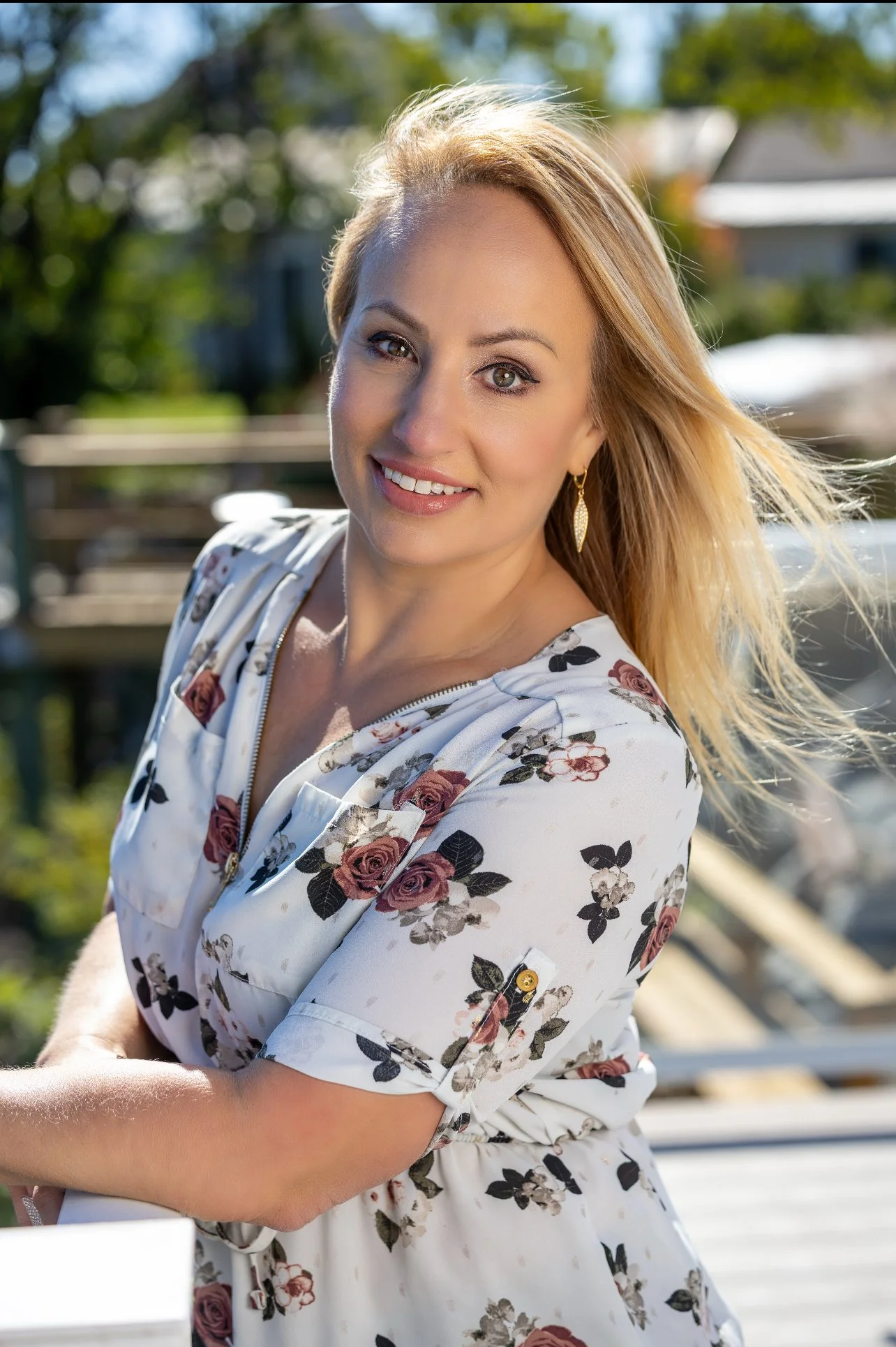 A woman with blonde, curly hair taking a selfie indoors, smiling, wearing a dark blue sleeveless top with a sheer neckline.