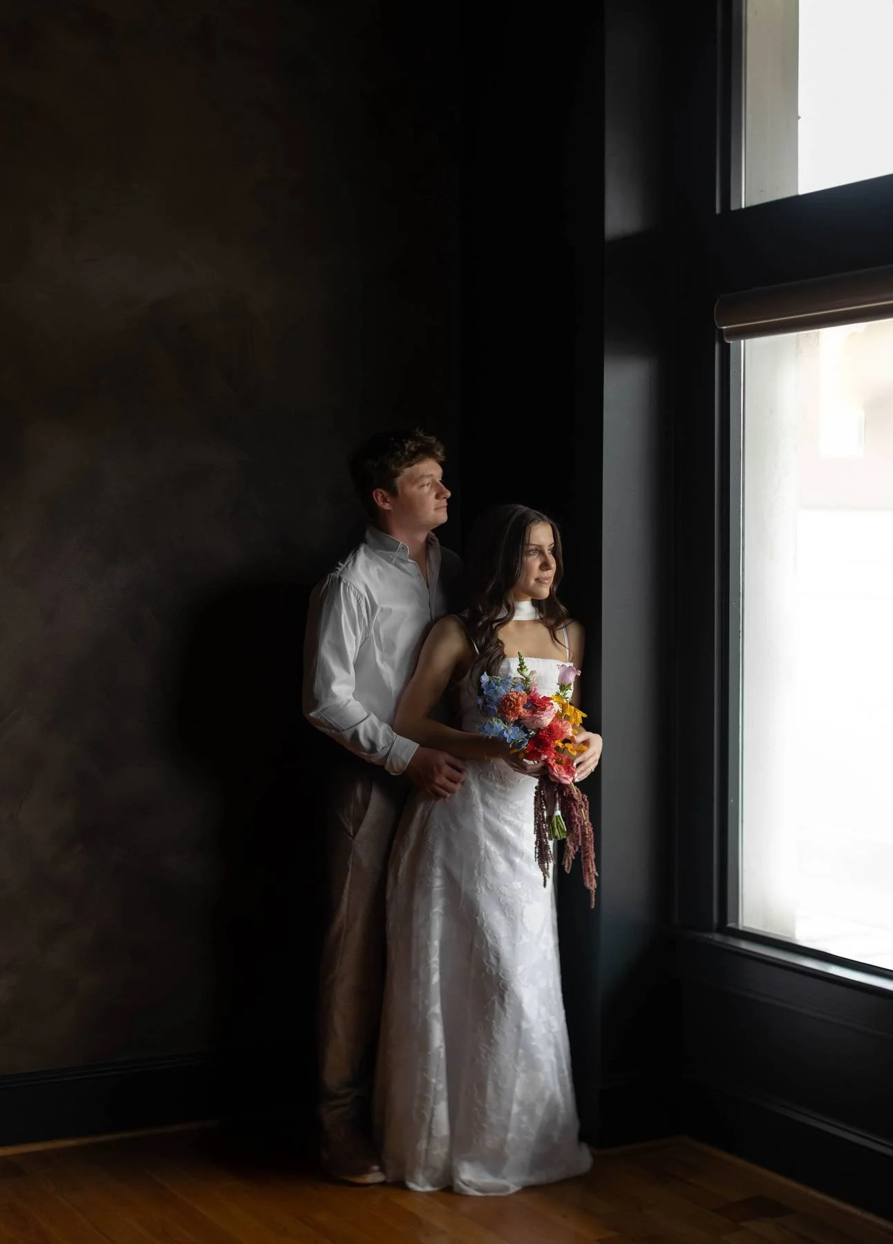 A couple in wedding attire standing by a large window, with the woman holding a bouquet of colorful flowers, gazing out contemplatively, in a dimly lit room with dark walls.
