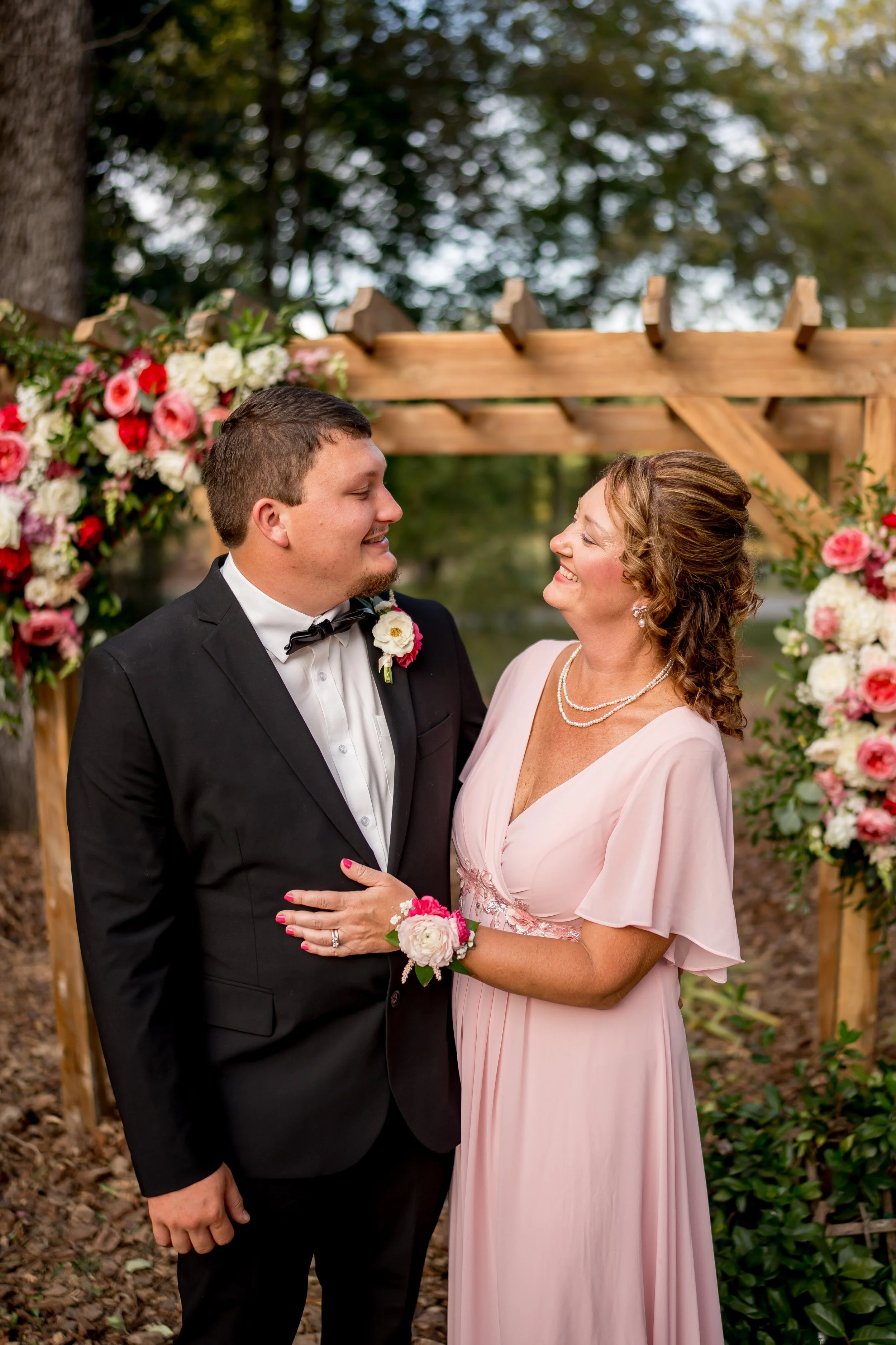 A bride and groom standing under a wooden arch decorated with pink and white flowers, smiling at each other during a wedding ceremony outdoors.