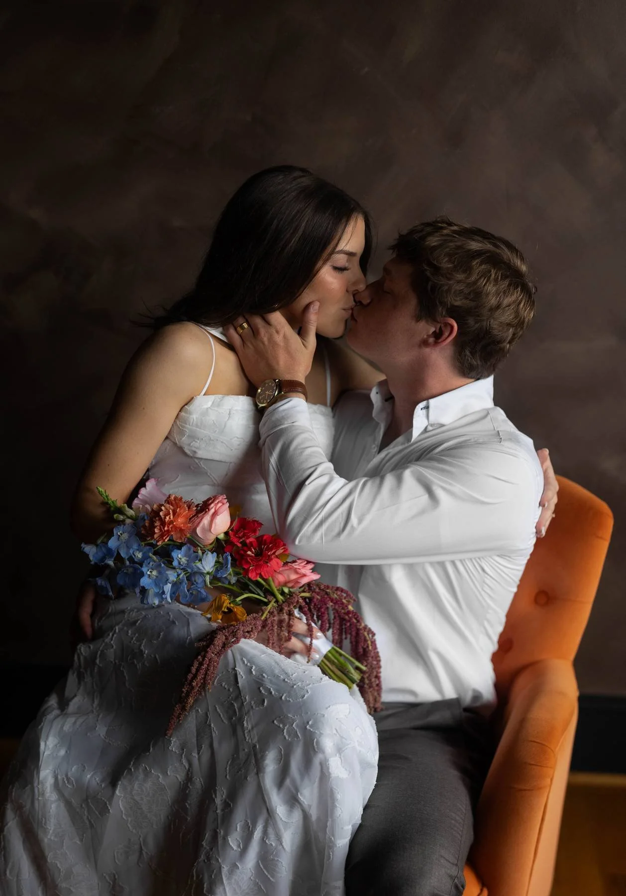 A man and woman share a kiss, with the woman sitting on a chair holding a bouquet of colorful flowers, and the man wearing a white shirt, seated beside her against a dark background.