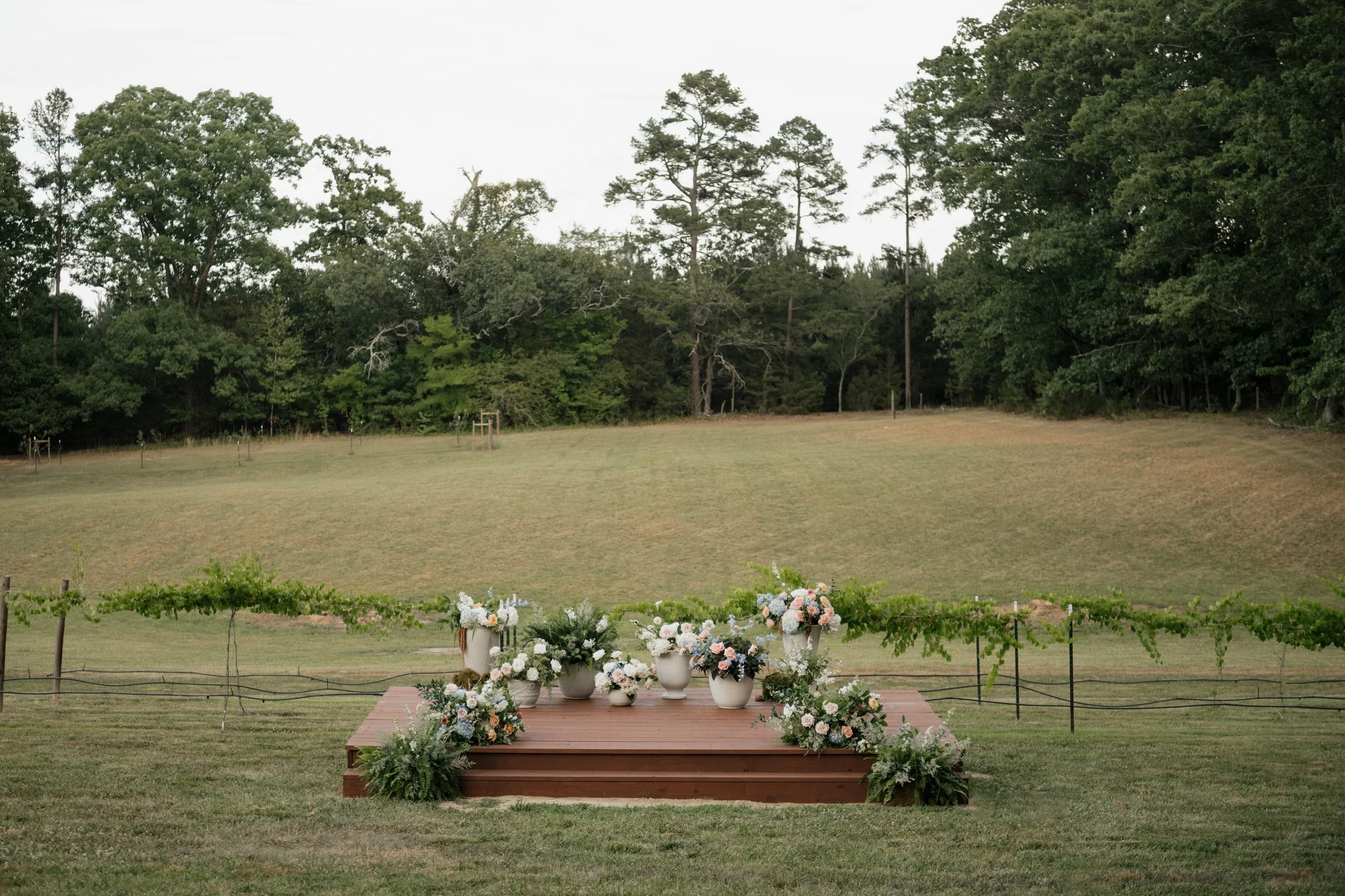 Beautiful outdoor wedding ceremony platform at wedding venue, surrounded by large floral arrangements in large open field and vineyard. Outside of Charlotte, North Carolina.
