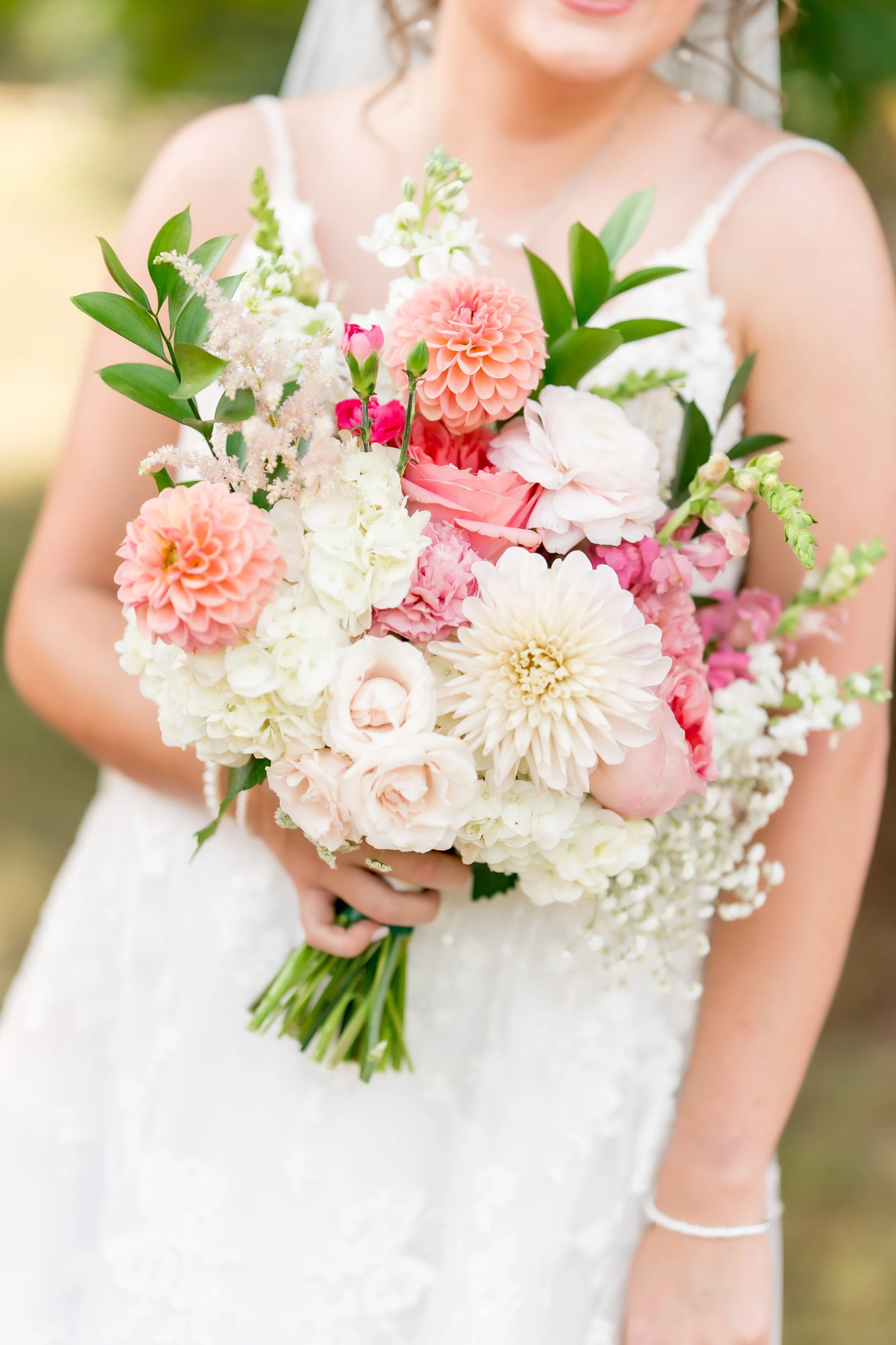 A woman in a white dress holding a large bouquet of pink, white, and cream flowers including dahlias, roses, and tulips, with greenery.