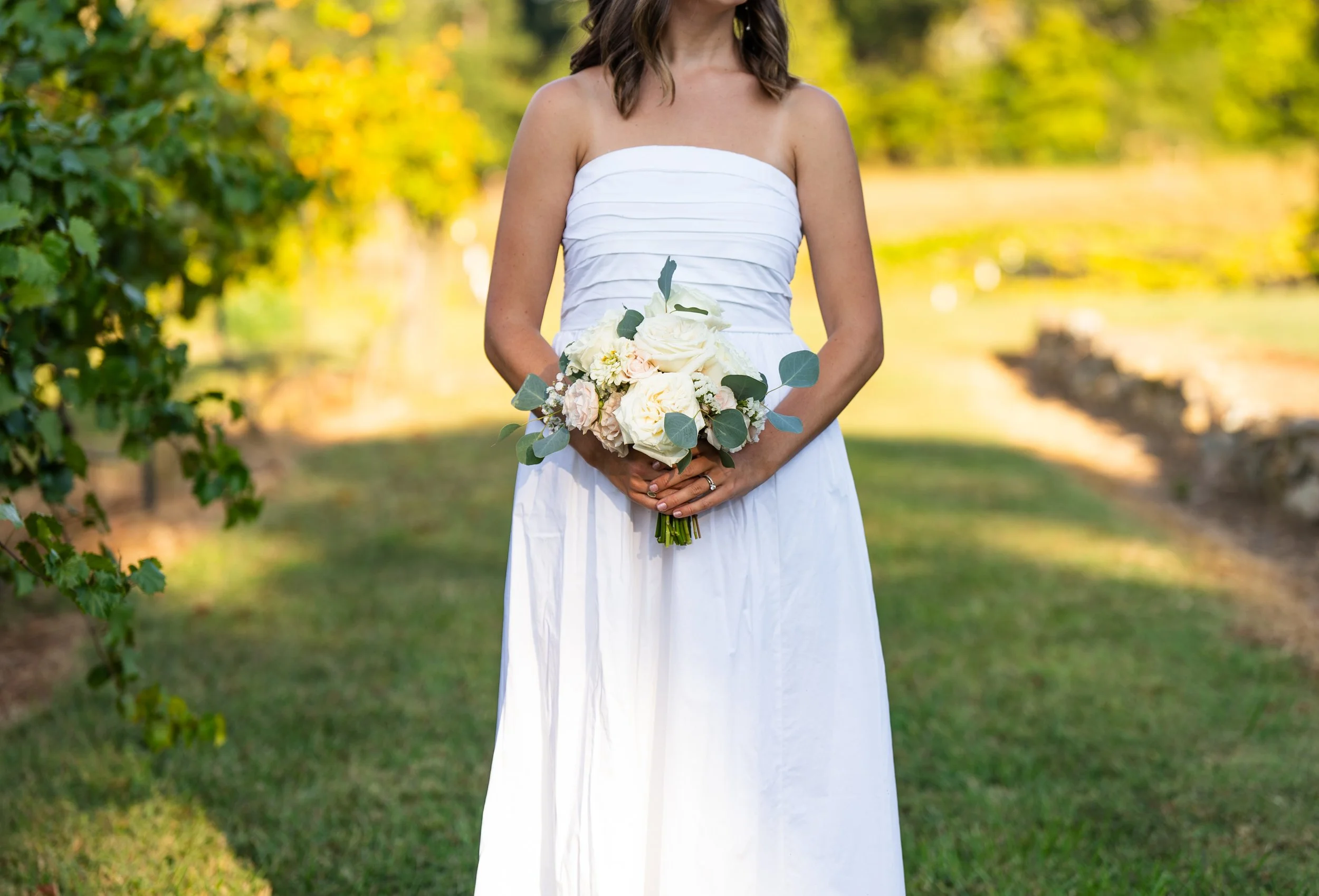 Woman in a strapless white dress holding a bouquet of white and blush flowers, standing outdoors on a grassy path surrounded by green trees.