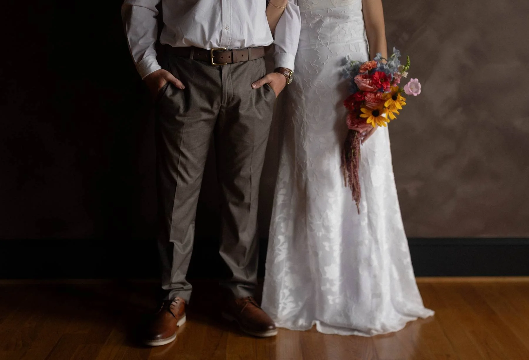 Partially visible couple, a man in dress pants, shirt, belt, and shoes, standing next to a woman in a wedding dress holding a colorful bouquet of flowers, against a dark background.