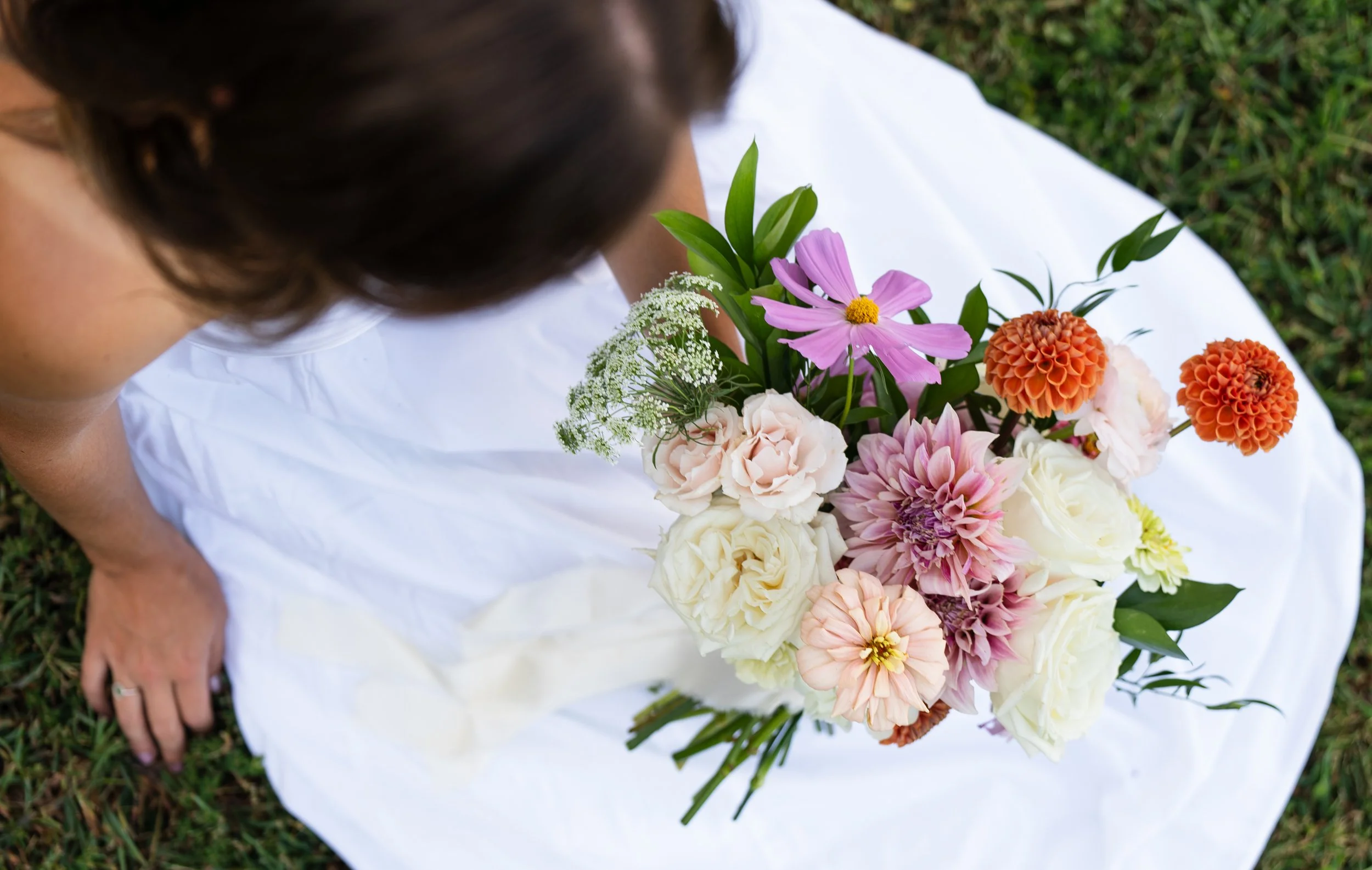 A bride sitting on the grass holding a bouquet of pink, white, and orange flowers, wearing a white dress. Wedding and elopement floral design.