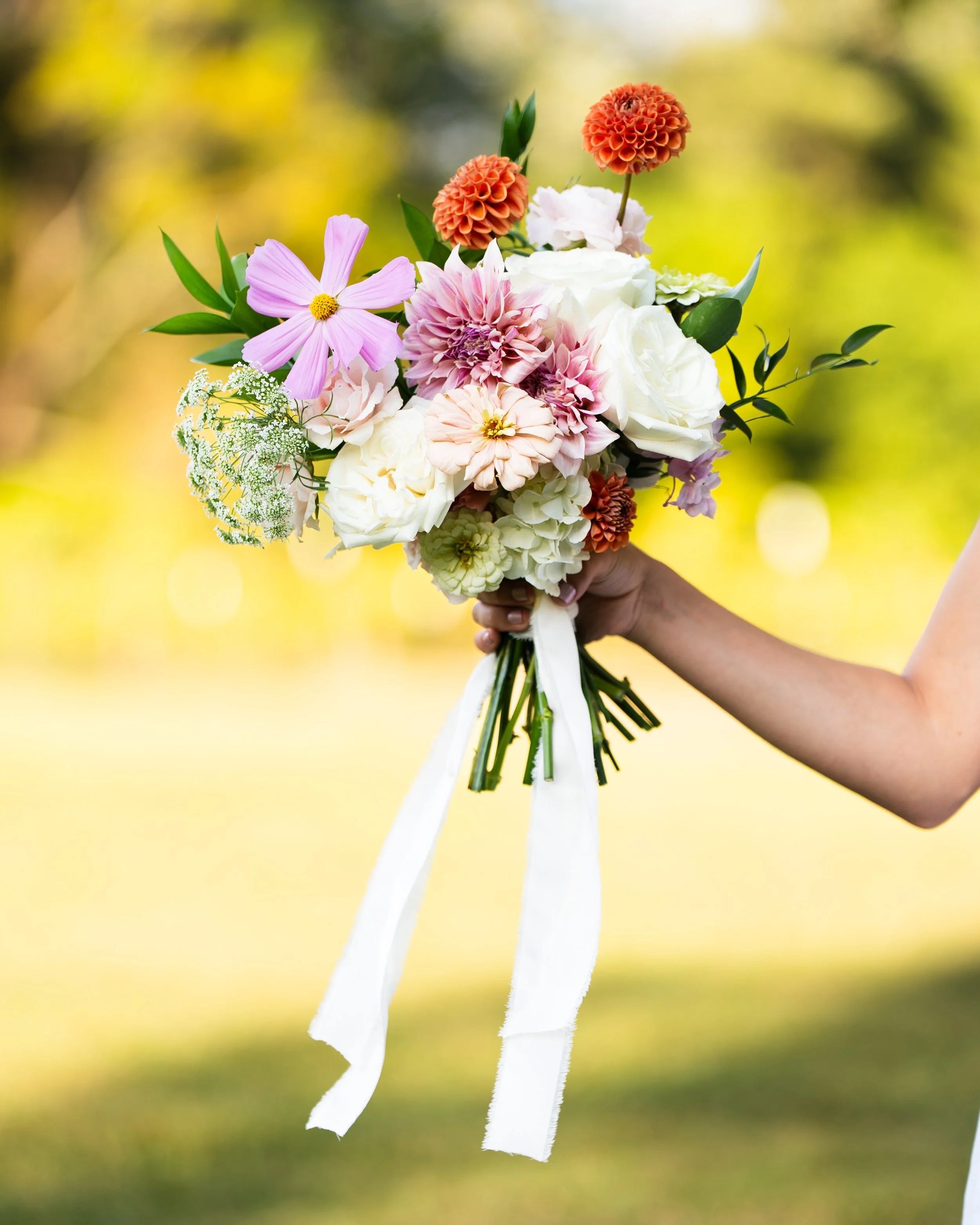 A hand holding a colorful bouquet of flowers, including pink, white, and orange blooms, with a white ribbon, outdoors on a blurred yellow and green background.