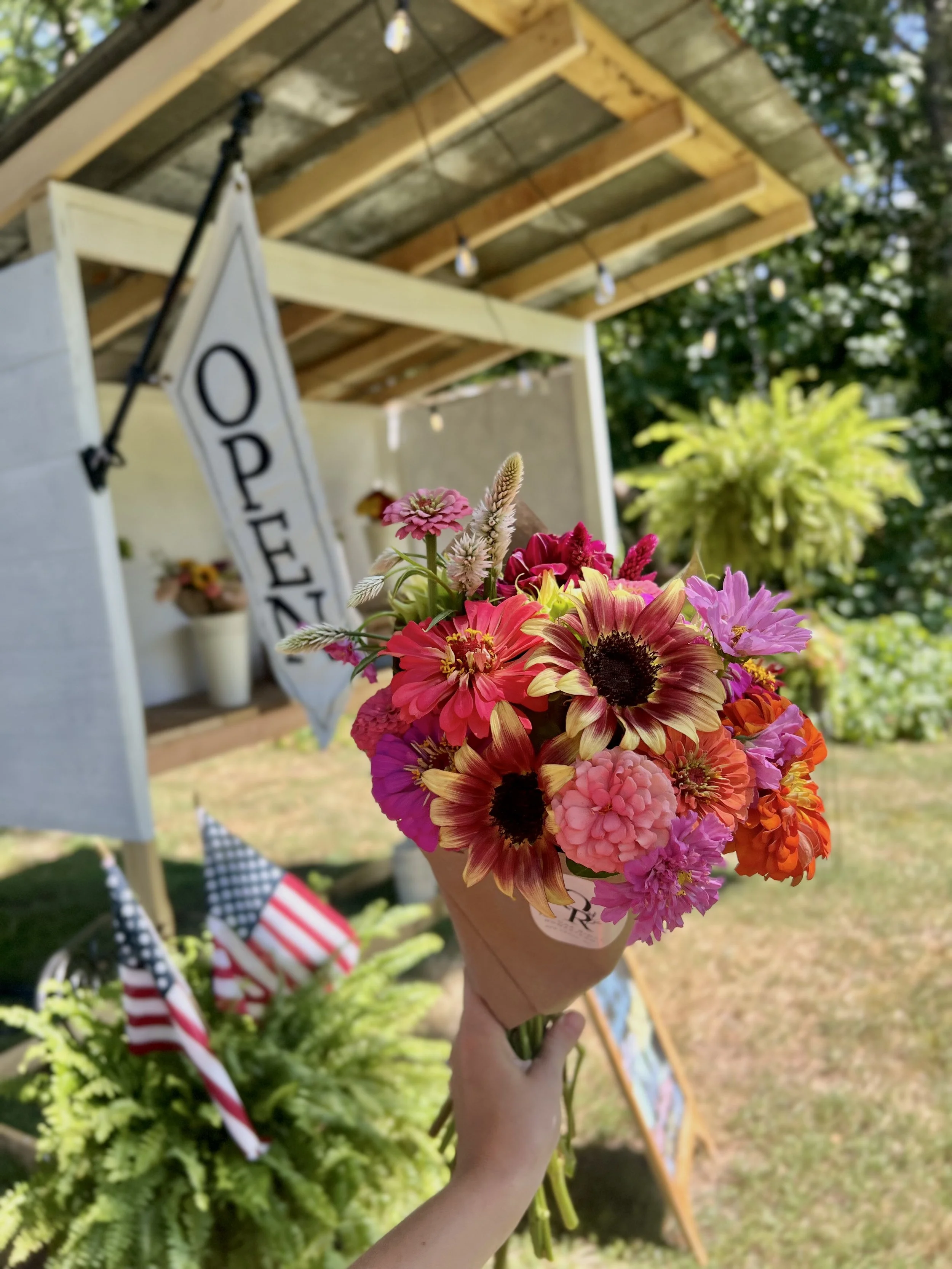 A hand holding a bouquet of colorful flowers, including pink, purple, orange, and yellow blooms, in an outdoor setting with a small wooden structure and American flags in the background. Flower and farm stand.