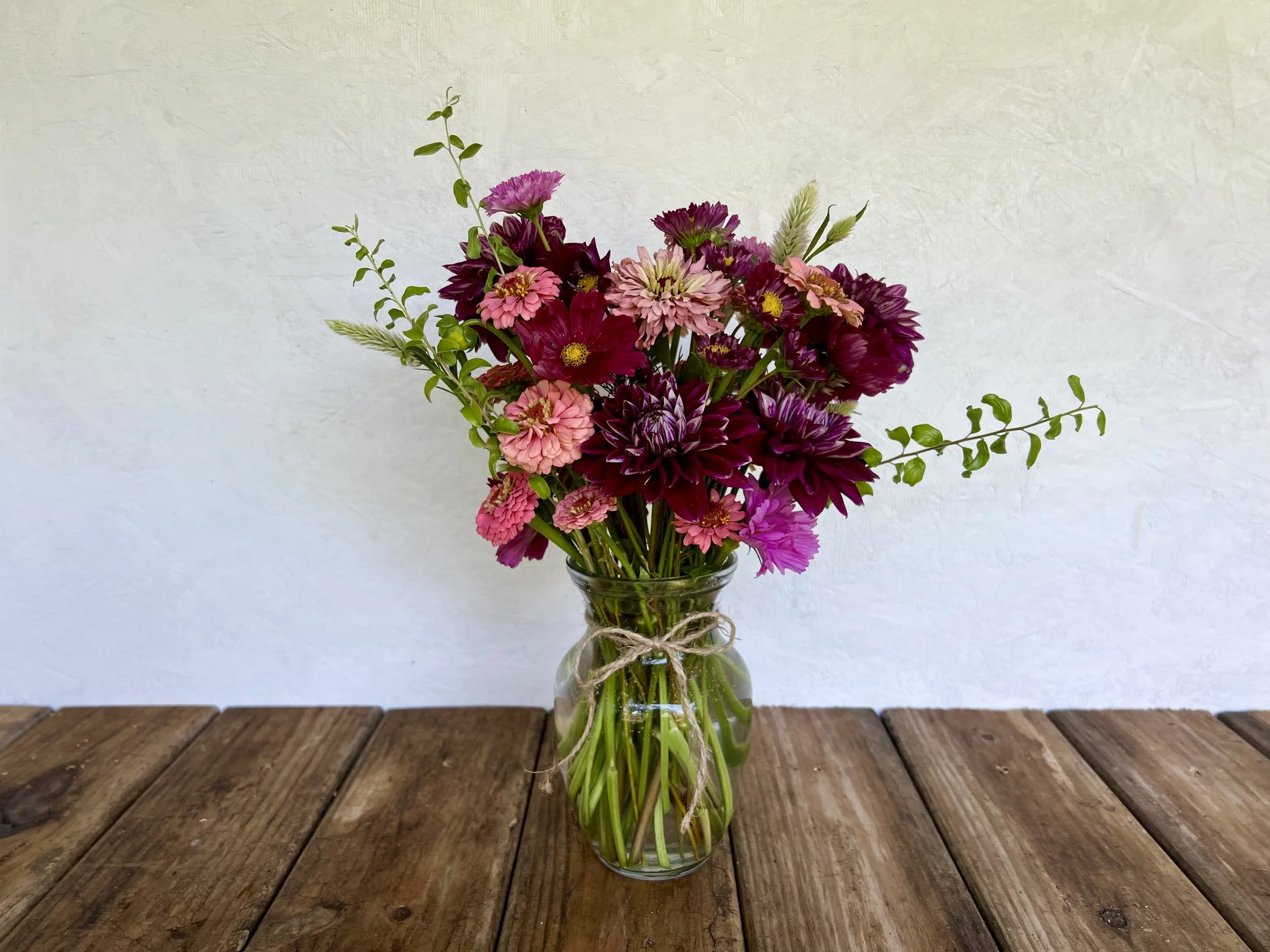 A glass vase filled with pink and purple dahlias on a wooden table against a plain wall.