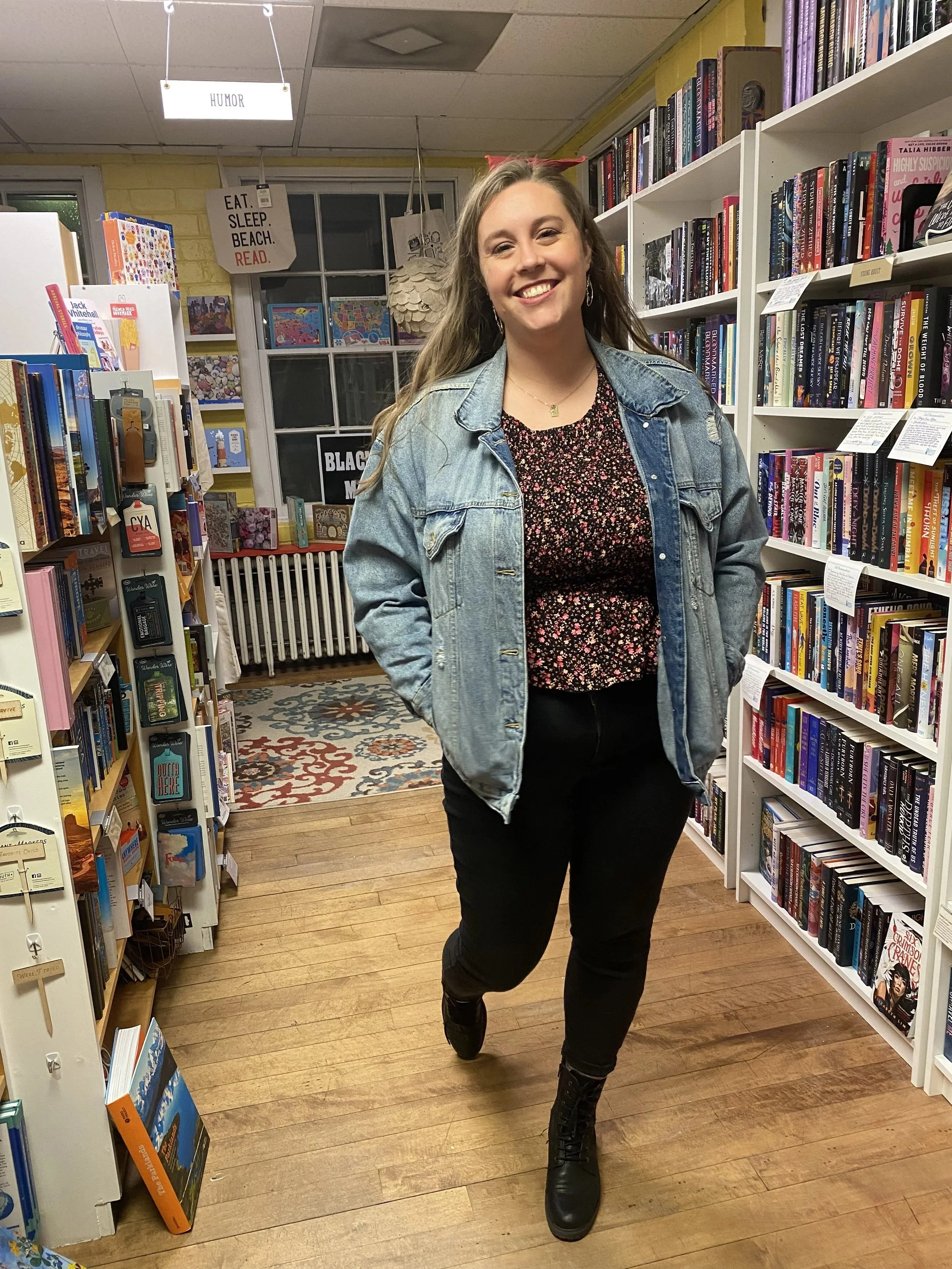 A woman smiling and walking through a bookstore with shelves of books on either side, wearing a denim jacket, floral top, black pants, and black boots.