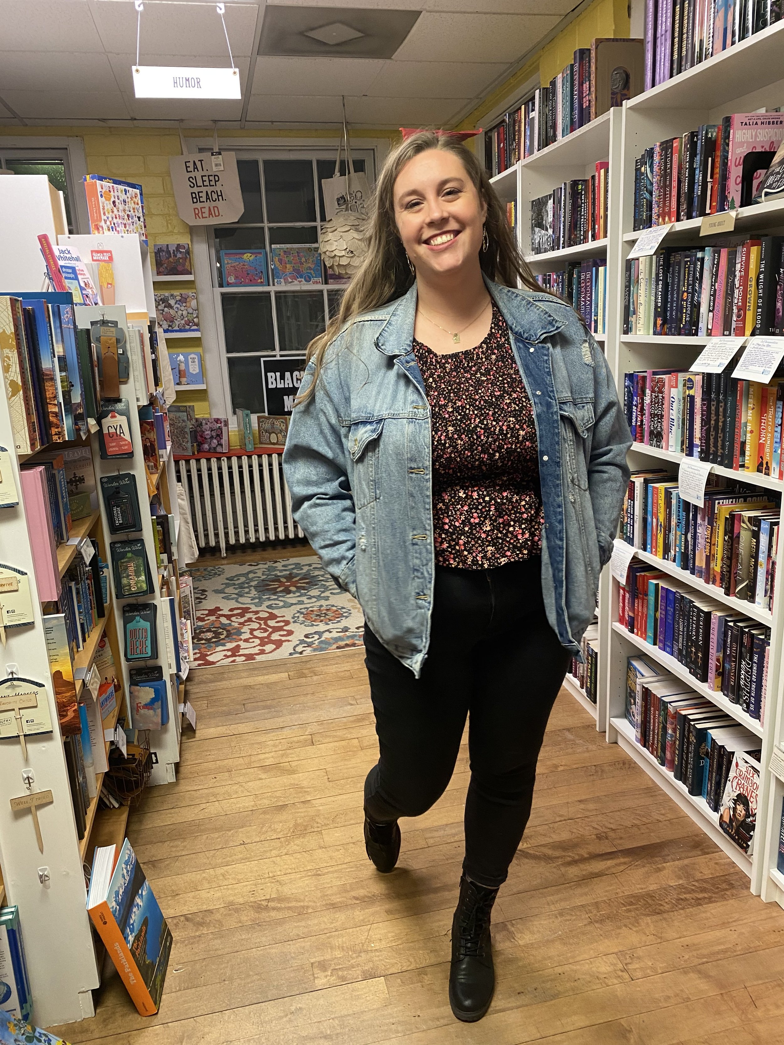 A woman with long blonde hair stands in a bookstore. She's wearing a jean jacket, floral top, black pants, and black boots.