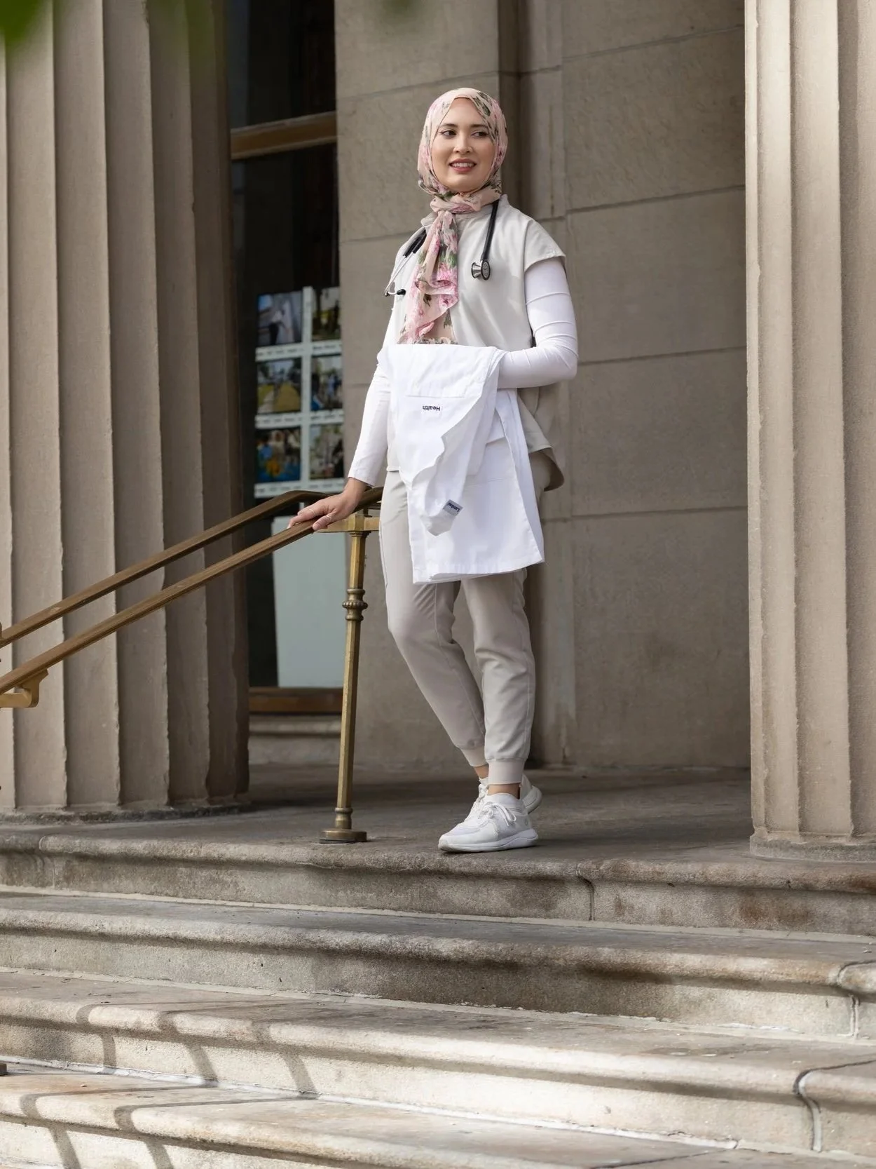 A female healthcare professional standing on steps outside a building, wearing a white coat, stethoscope, and a floral headscarf, holding a white shirt.