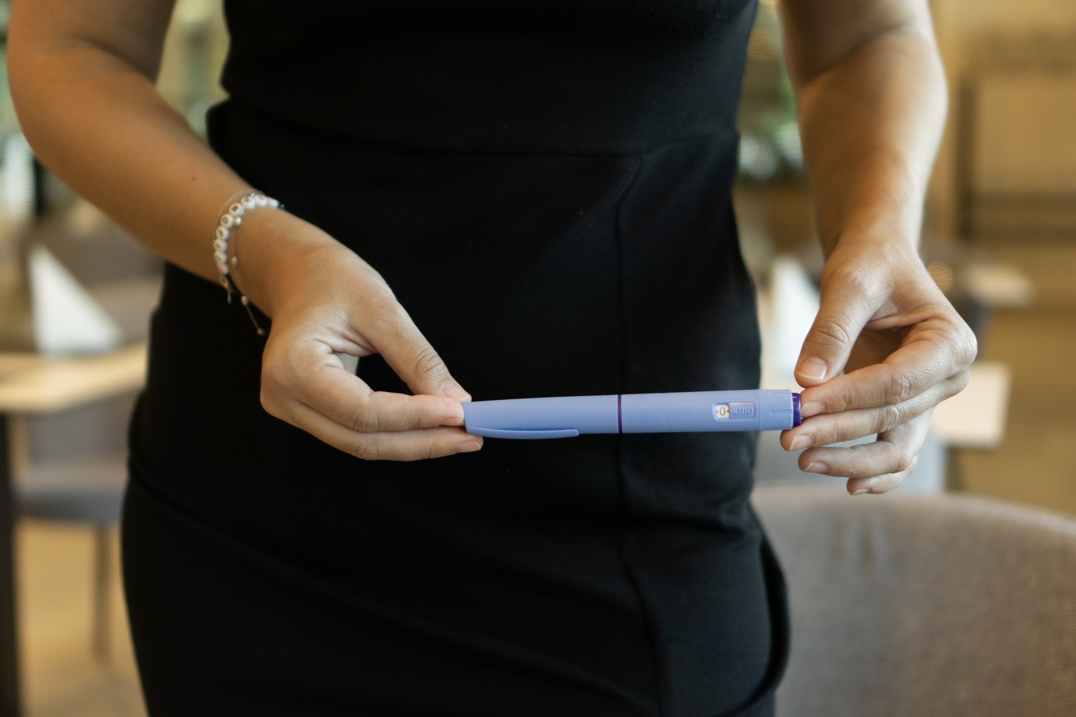 Person holding a blue insulin pen with a small window displaying '0' and 'mg' in a room with wooden furniture.