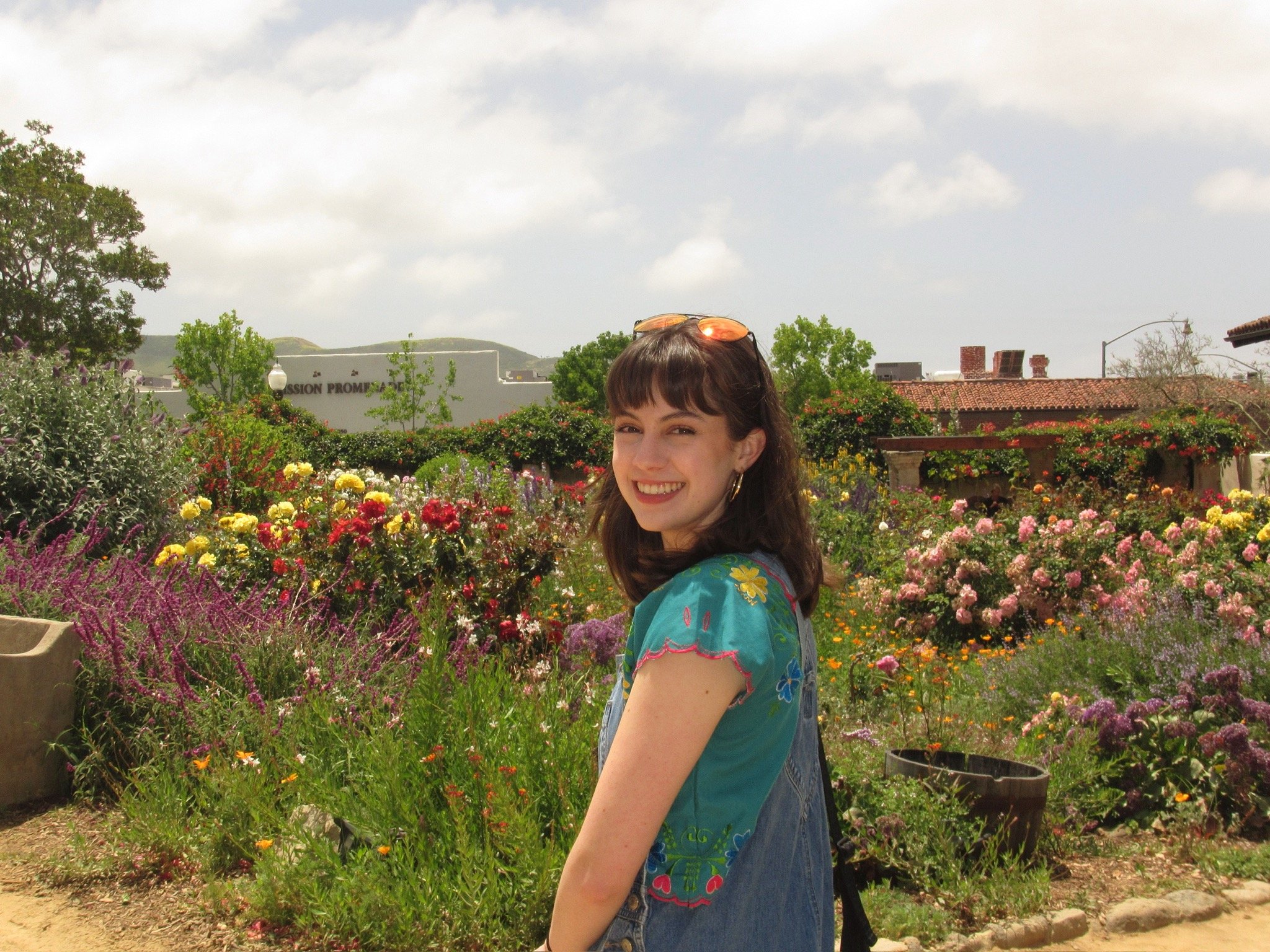 Gabriela Ayala Yarbrough standing in front of flowers turning over her shoulder and smiling at the camera wearing a green floral top and overalls