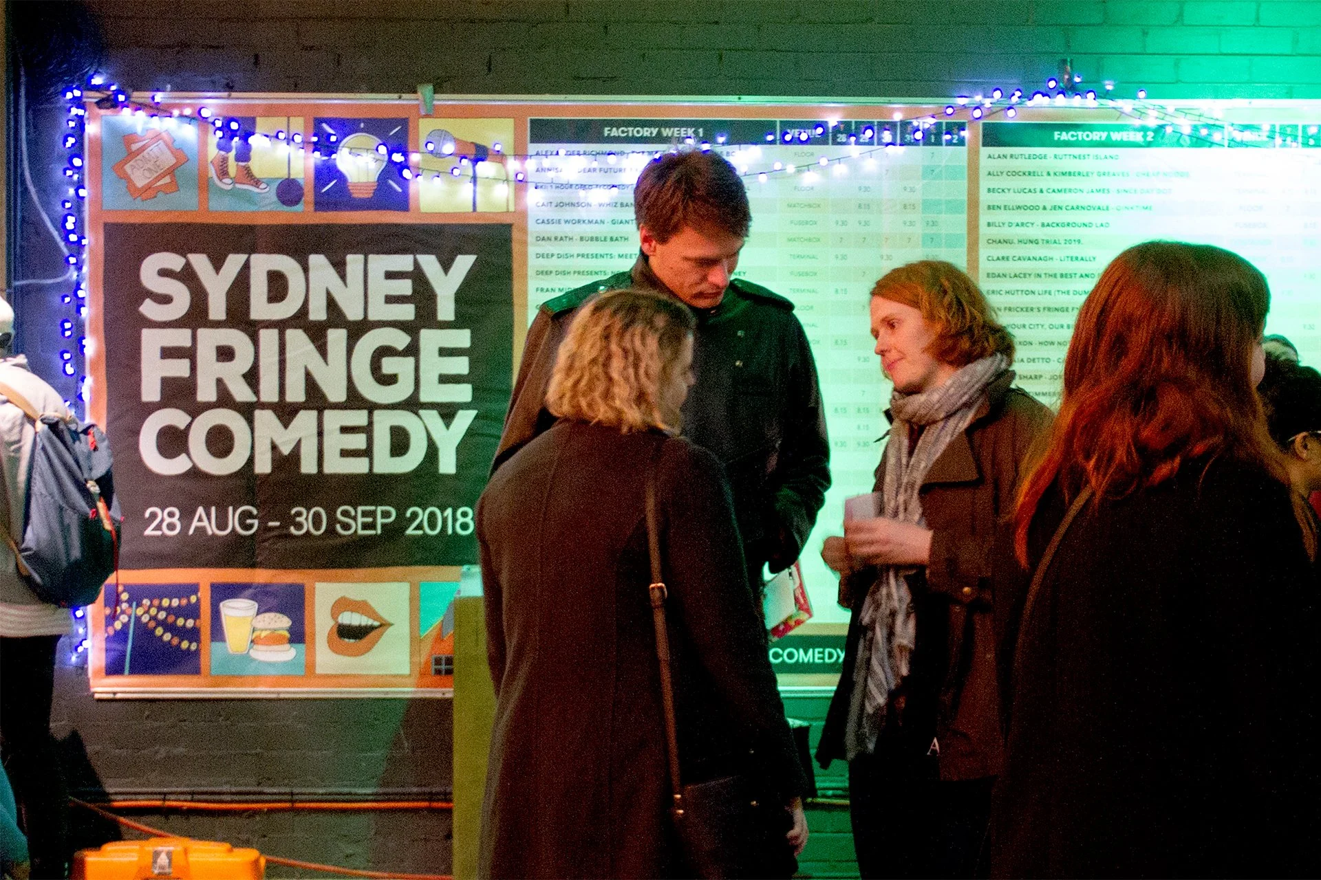 Group of people talking outside at the Sydney Fringe Comedy event, with a large schedule board and colorful lights in the background.