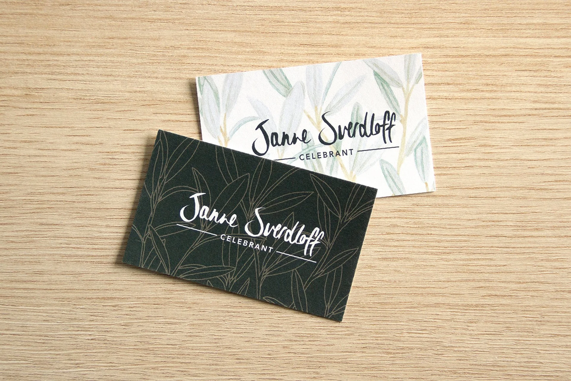 Two business cards for Jeanne Svedoff, one with a white background and green leaf pattern, the other with a dark background and gold leaf outline pattern, both on a wooden surface.