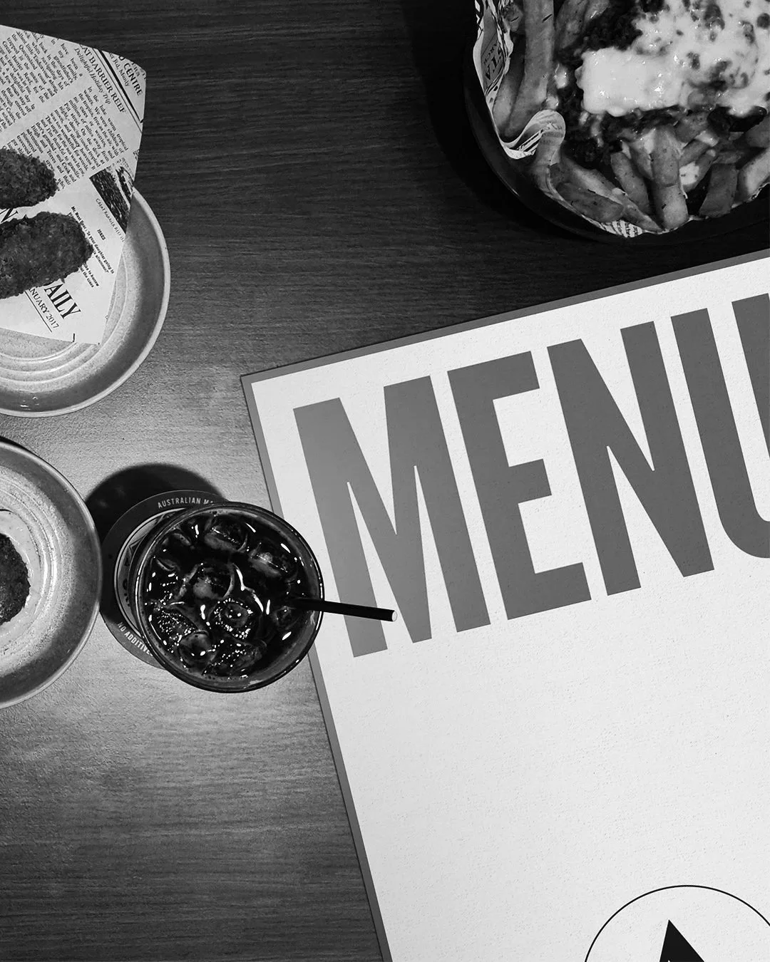 A restaurant table with a menu, a bowl of French fries topped with cheese and bacon, a small plate with fried food, and a glass of soda with ice and a straw.