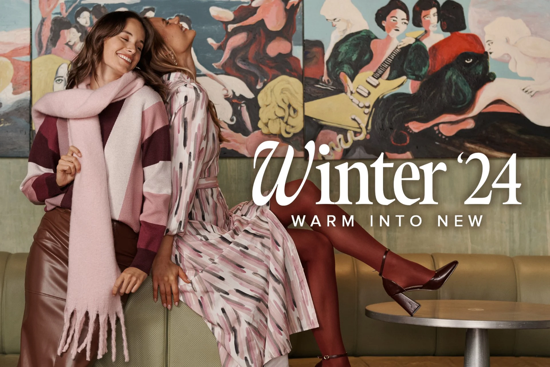 Two women smiling and leaning on each other in a cafe, with colorful artwork on the wall behind them; one woman sits on a bench with her legs crossed and wears a striped dress and maroon heels; the other stands beside her, wearing a patterned sweater, a scarf, and leather skirt.