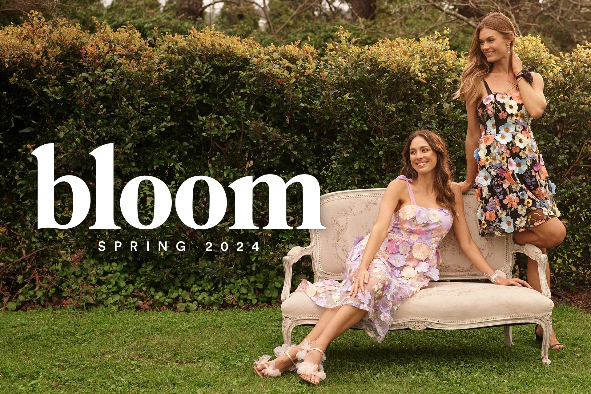 Two women wearing floral dresses posing on a vintage sofa outdoors, with a bush and trees in the background, promoting Bloom Spring 2024 collection.