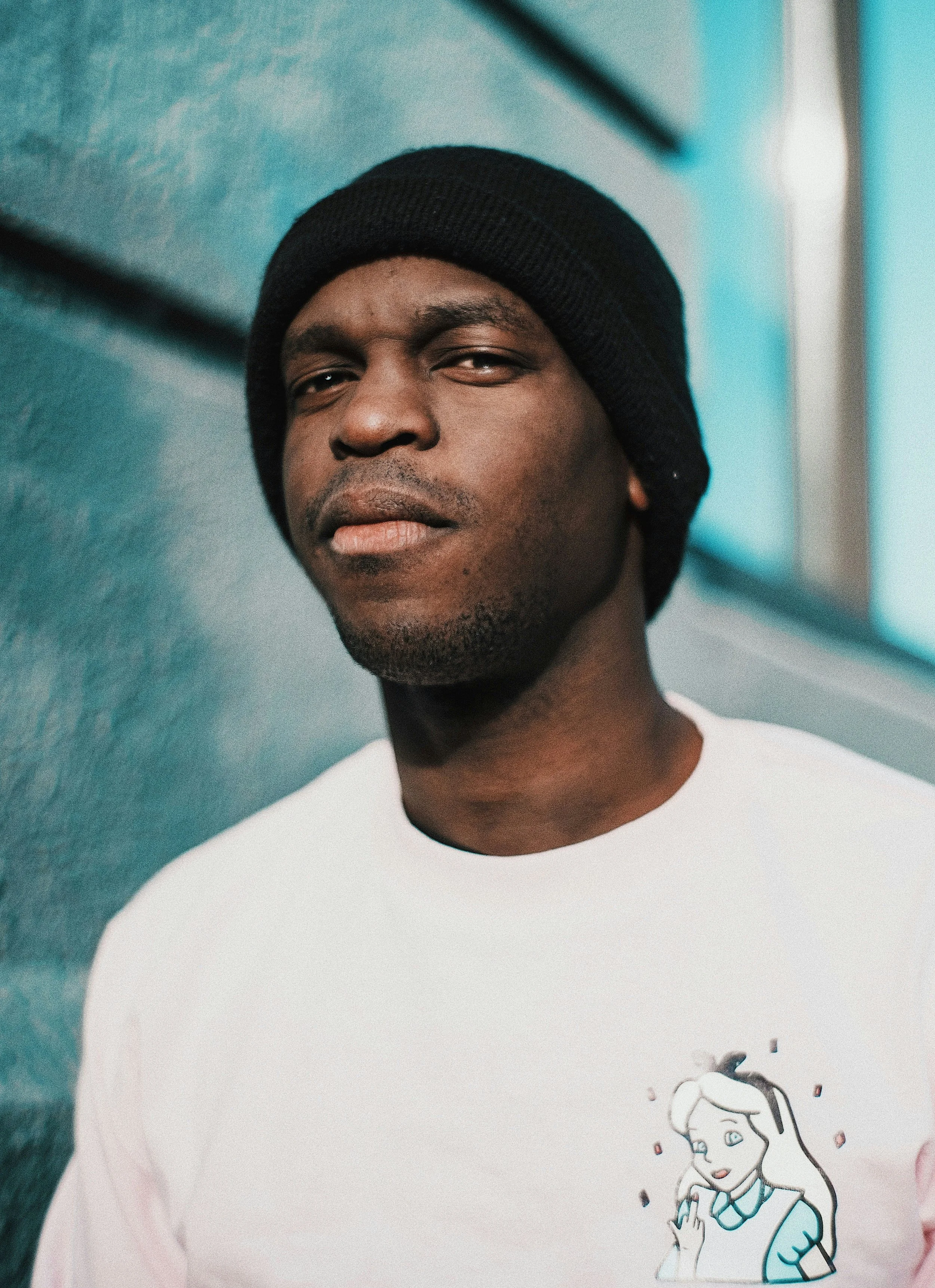 A young man wearing a black beanie and a white t-shirt with a cartoon girl on it, standing against a blue wall.