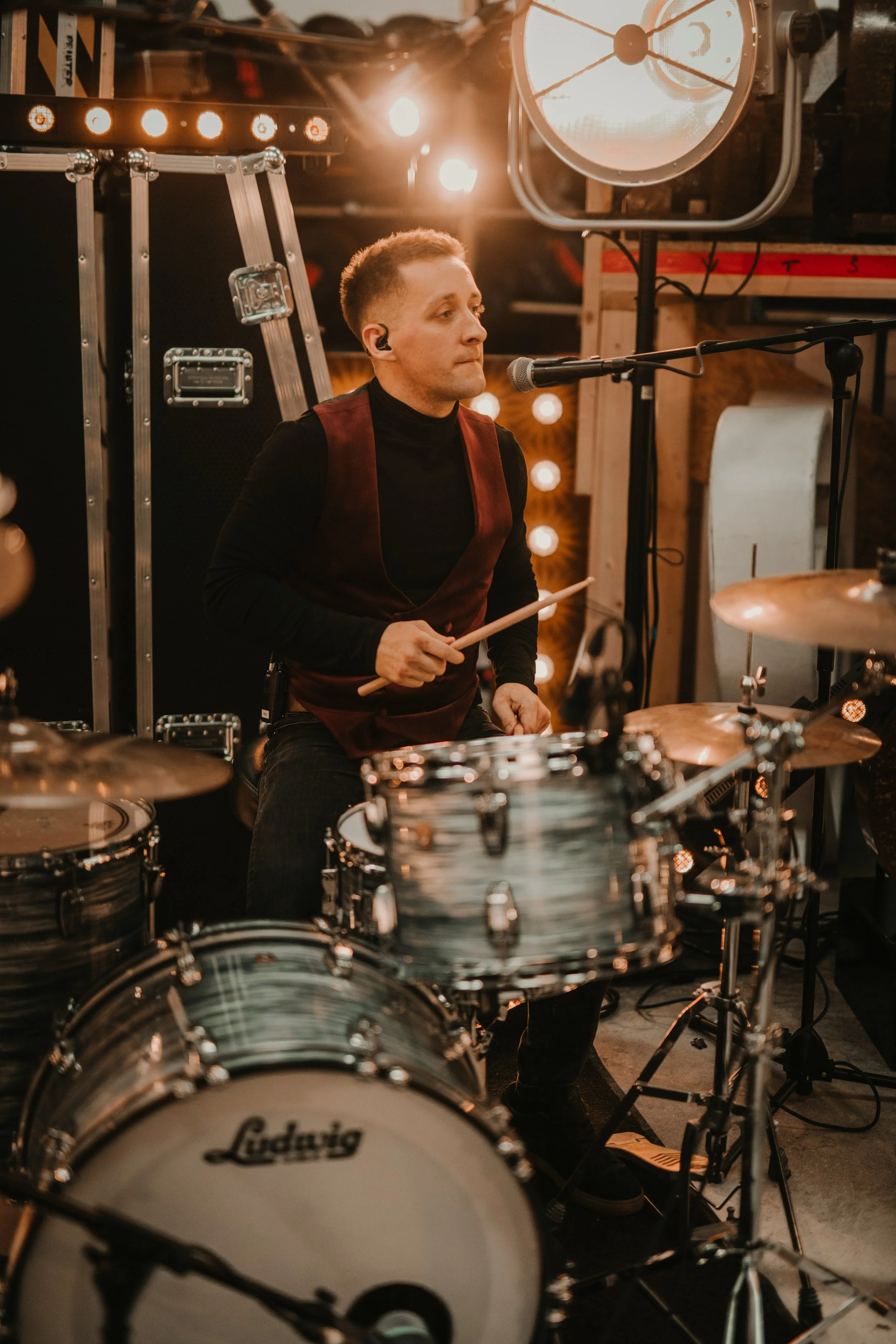 A male drummer playing a drum set during a live performance in a studio with warm lighting.