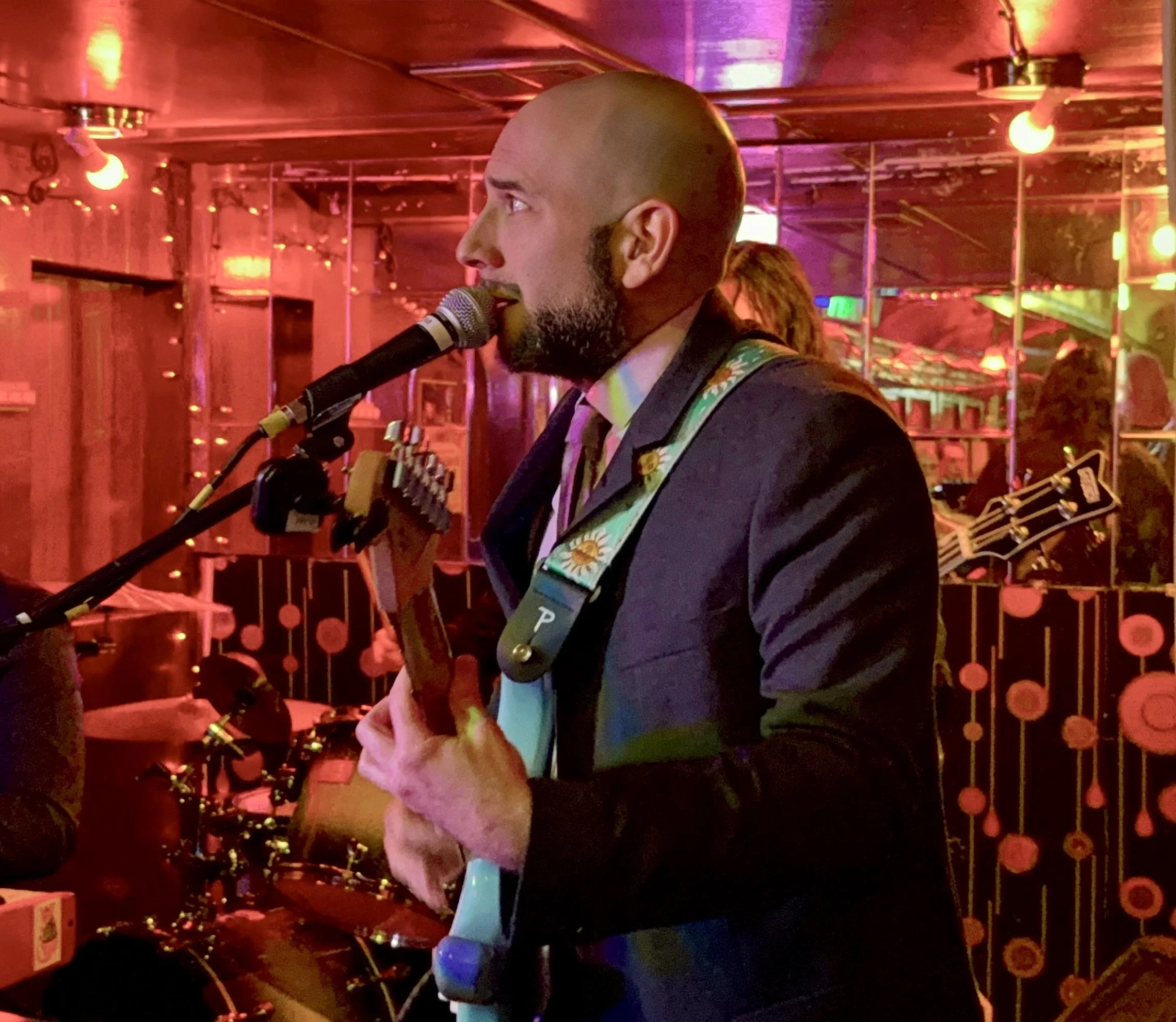 Jonathan Flom in a suit singing into a microphone in a dimly lit bar or club with warm lighting, people in the background, and decorative string lights.