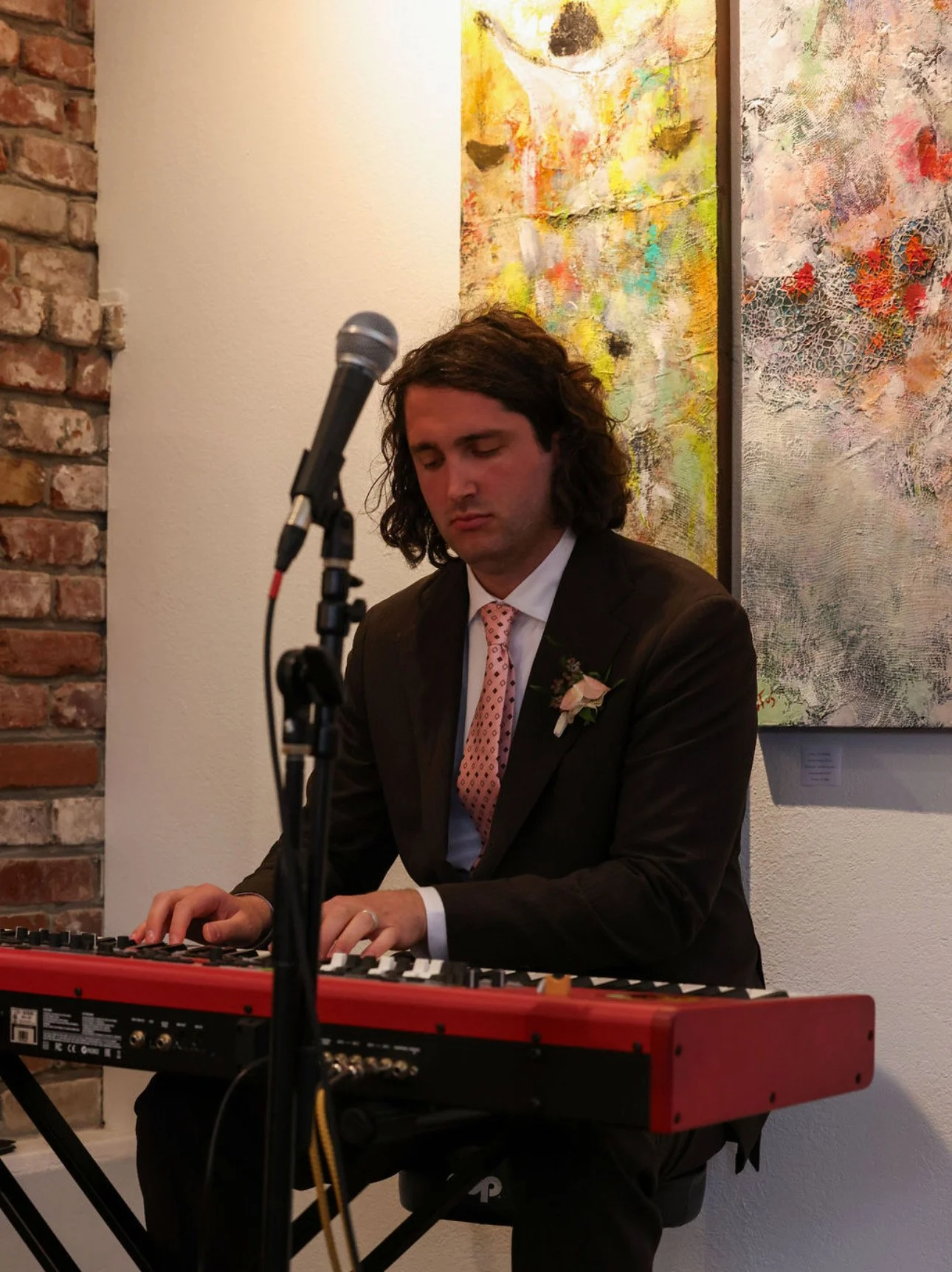 Jacob Cleveland in a suit with a pink tie and a boutonniere playing a red keyboard at an art gallery with abstract paintings on the wall behind him.
