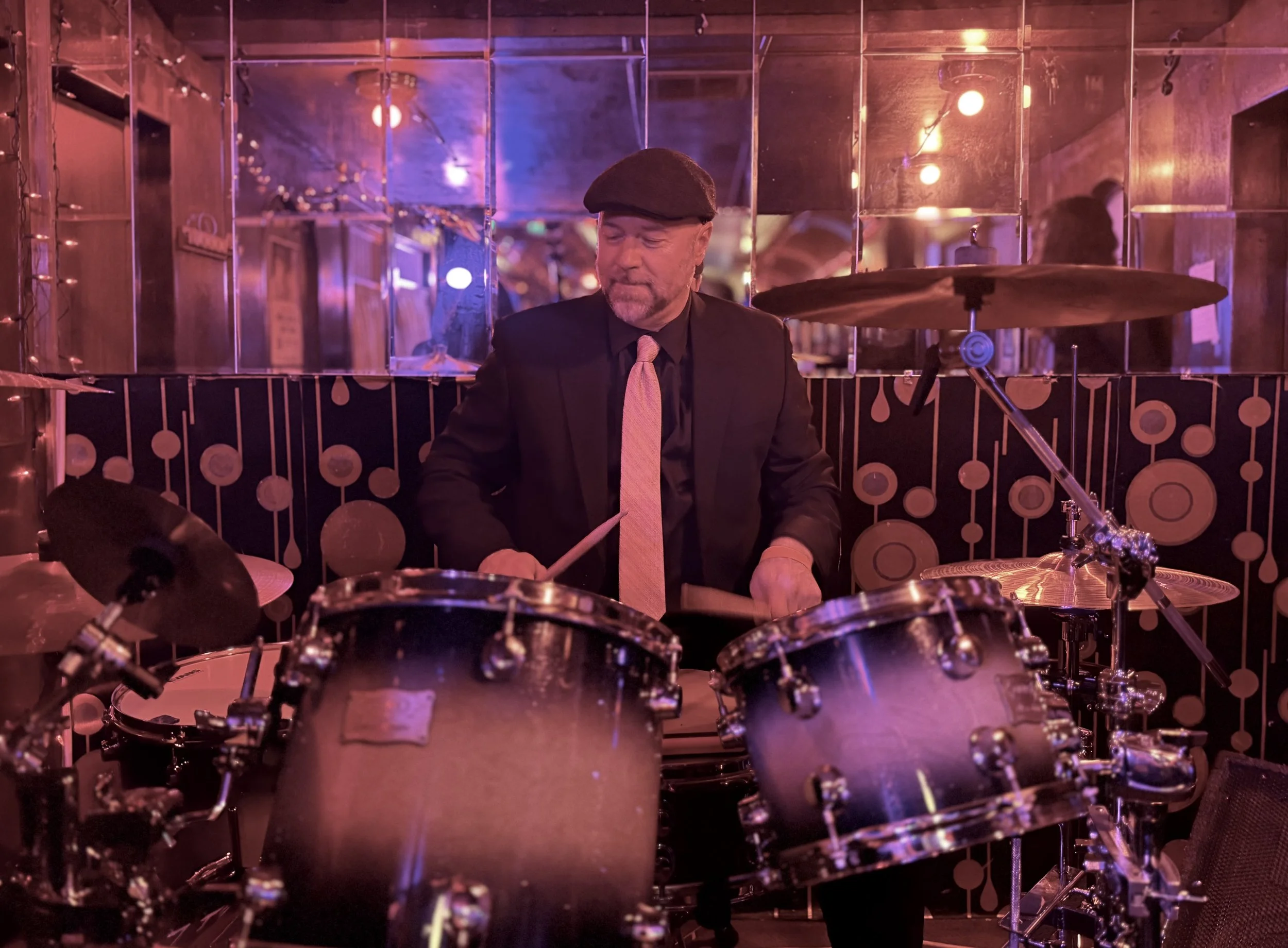 Peter Berger in a black suit, pink tie, and brown hat playing drums in a dimly lit room with pink and purple lighting, mirrored walls, and circular decorations.