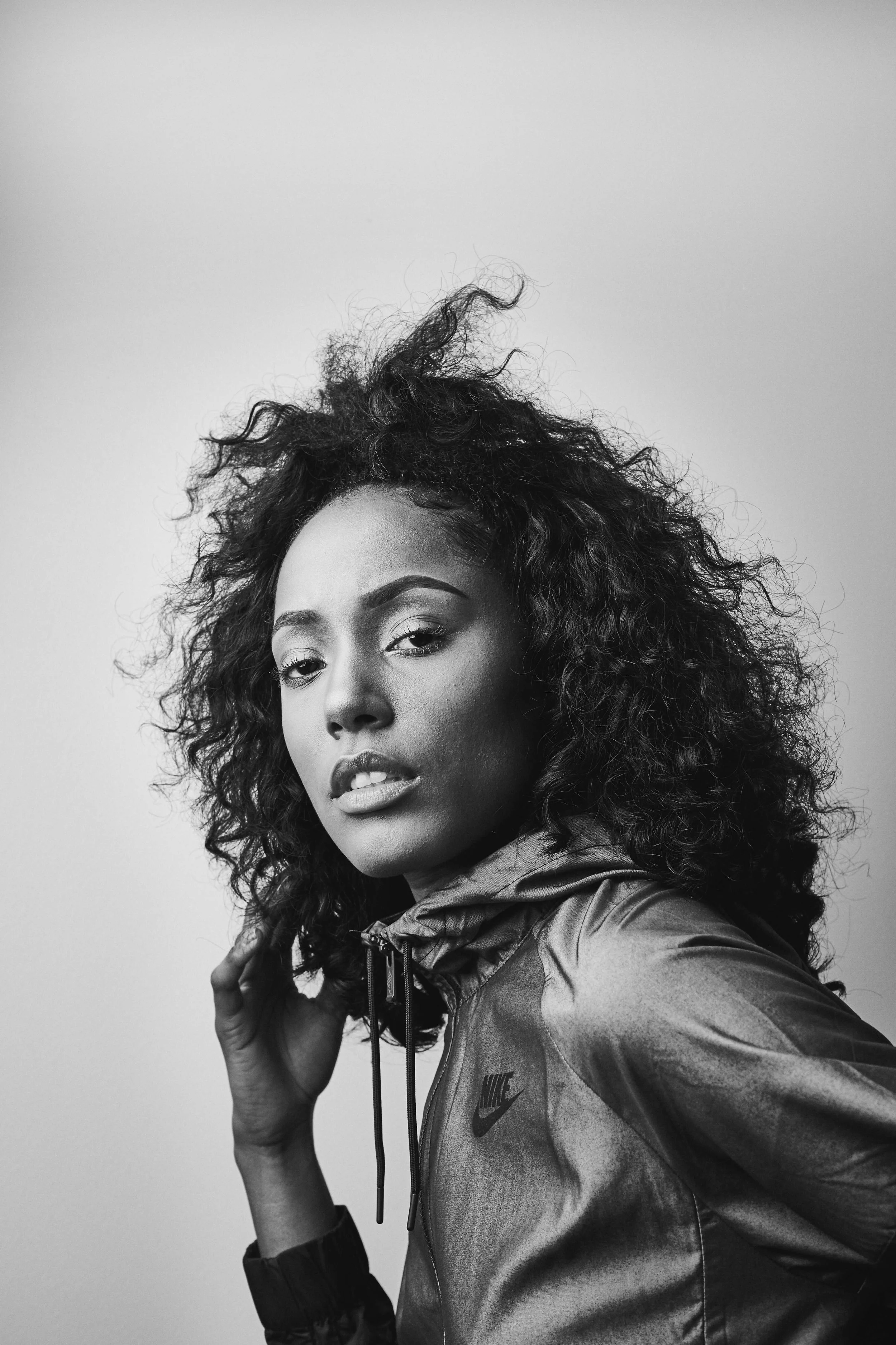 Black and white portrait of a woman with curly hair wearing a Nike jacket, looking at the camera.