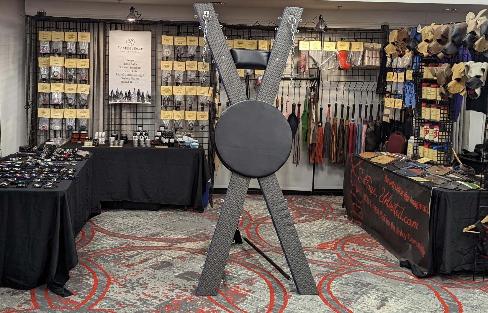 Display booth at a trade show with various leather accessories. Centered is a large, black, circular padded pad attached to a metal structure. On the left, tables with small leather items. On the right, tables with accessories. The back wall has a grid with shelves filled with bottles, boxes, and a sign with the logo 'LeatherBear' listing products like soaps, bath salts, and beard balms.
