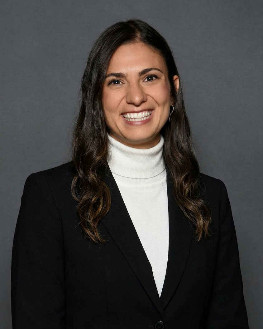 A woman with dark brown wavy hair wearing a black blazer over a white turtleneck, smiling against a gray background.