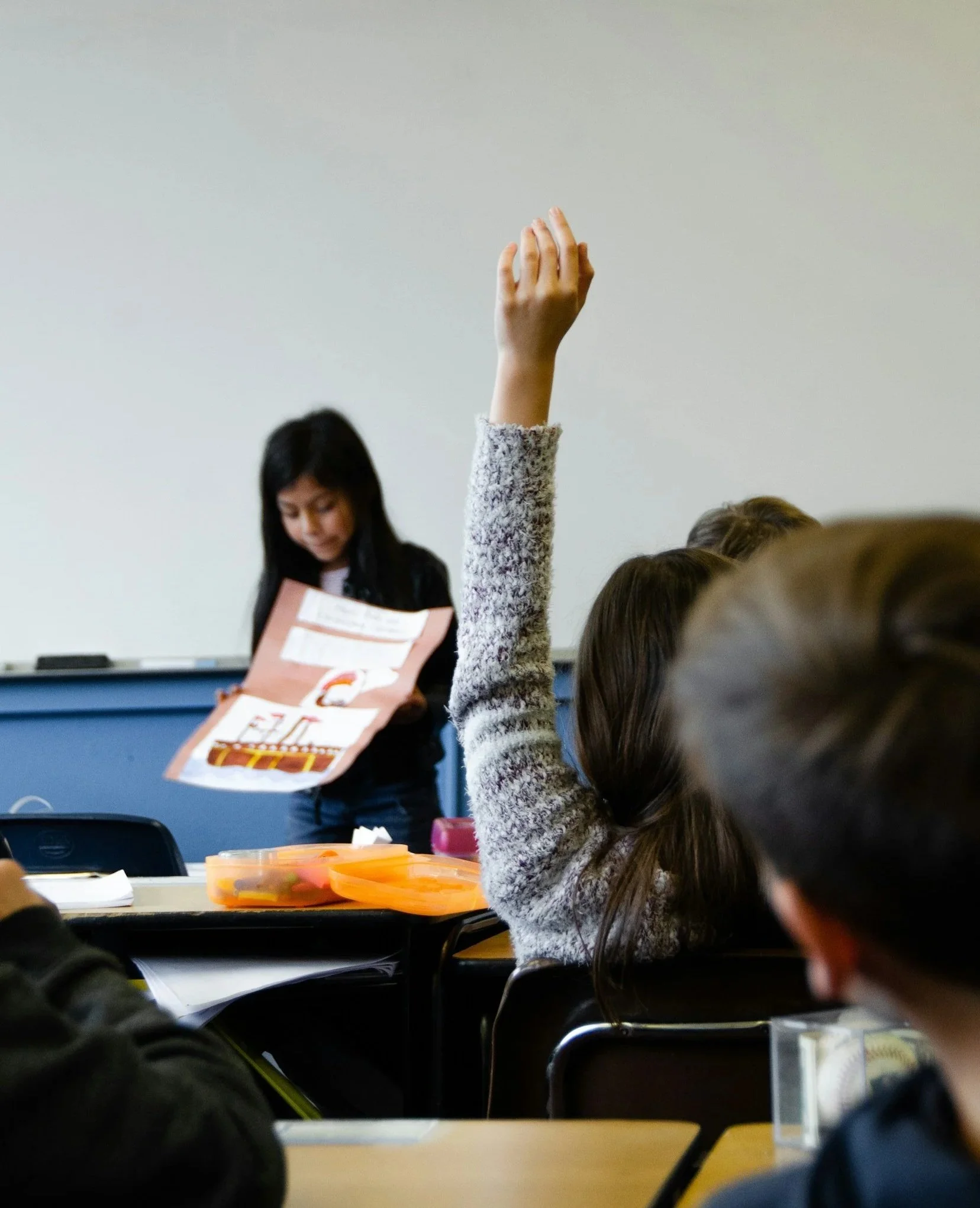 A classroom with students, one girl in the background holding a poster, and a student in the foreground raising their hand.