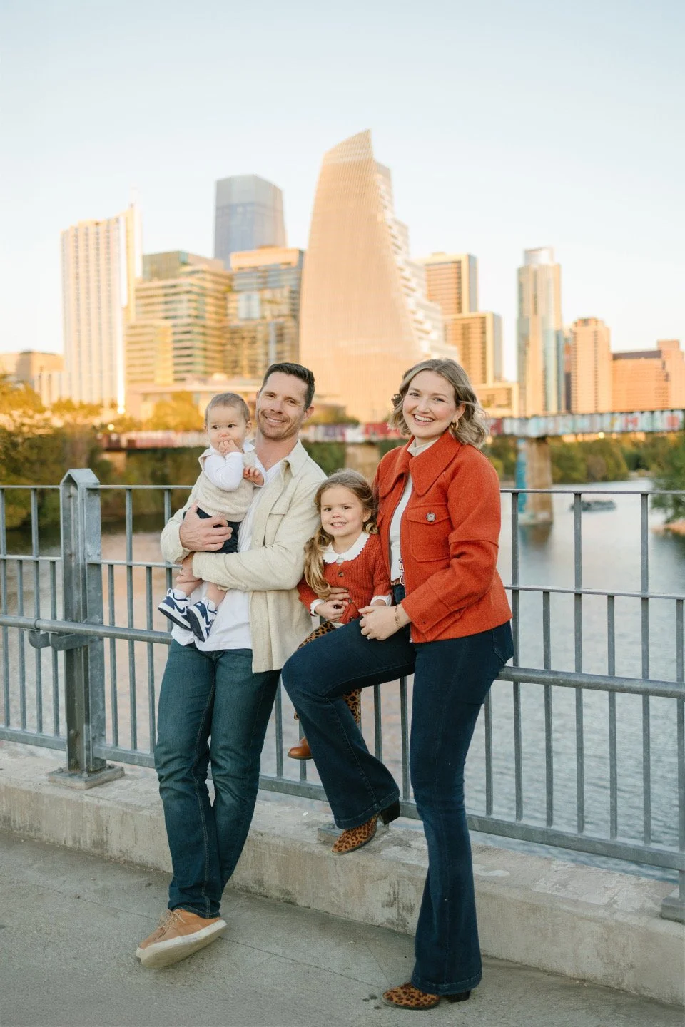 A family of four posing on a bridge with a city skyline in the background during sunset.