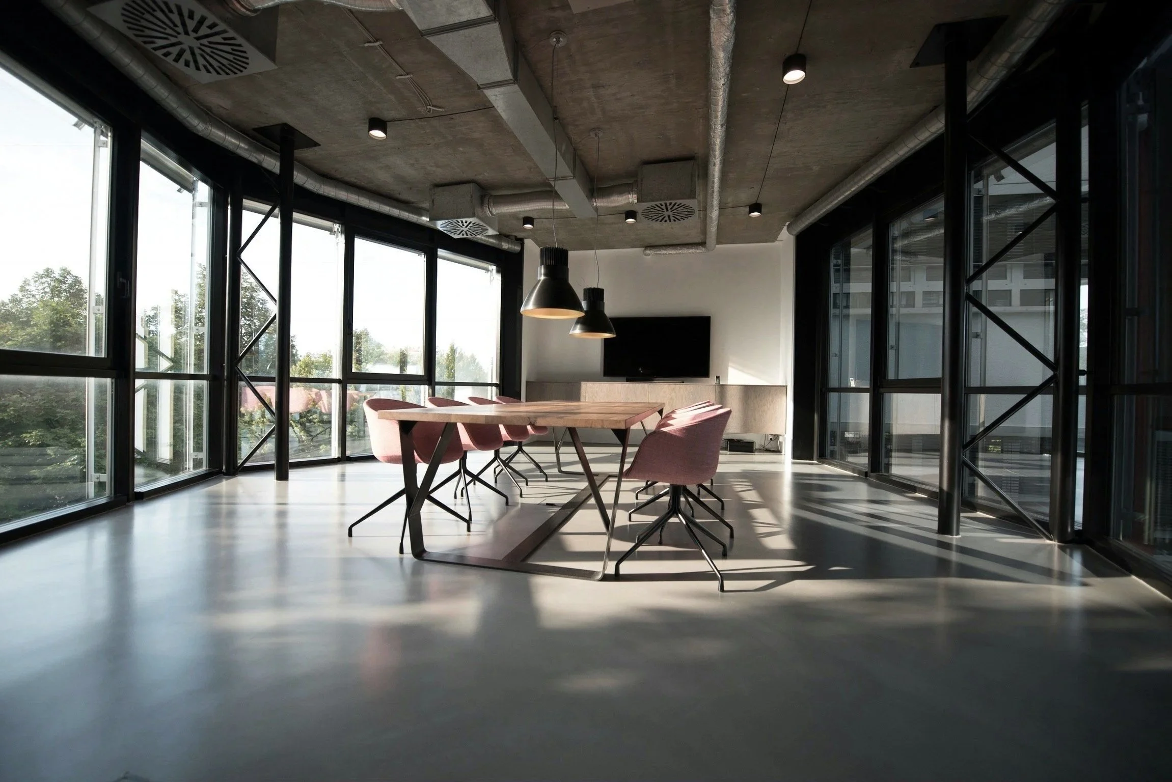 Modern conference room with large windows, pink chairs, wooden table, black hanging lamps, and a television on the back wall.