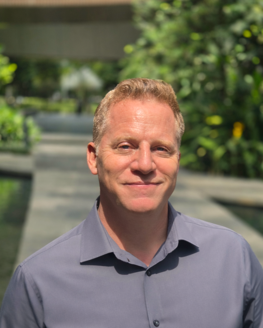 A man with short, light brown hair and wearing a gray button-up shirt, standing outdoors with greenery in the background.